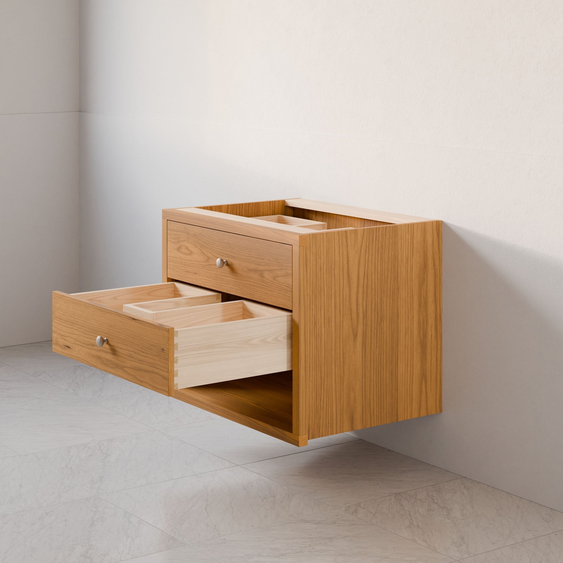 The Double Drawer, Open Shelf Vanity in Cherry by Krovel Furniture Co. is beautifully set against a light wall, featuring a hardwood veneer with a natural wood grain finish, sleek silver knobs, and dovetail joinery. It is elegantly mounted above a light-colored marble floor.