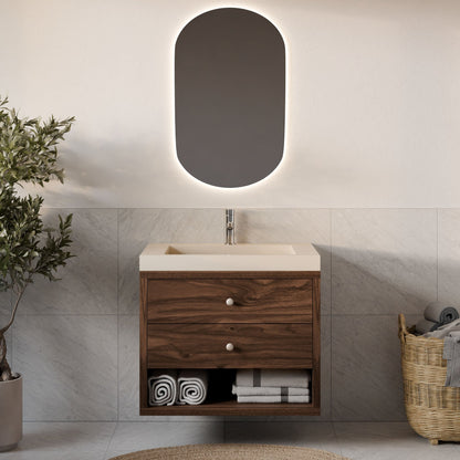 A contemporary bathroom features Krovel Furniture Co.'s Double Drawer, Open Shelf Vanity in Walnut, complemented by a custom-sized sink. An oval backlit mirror is mounted above. Rolled towels and toiletries are organized beneath the vanity, with a wicker basket and a potted plant nearby.