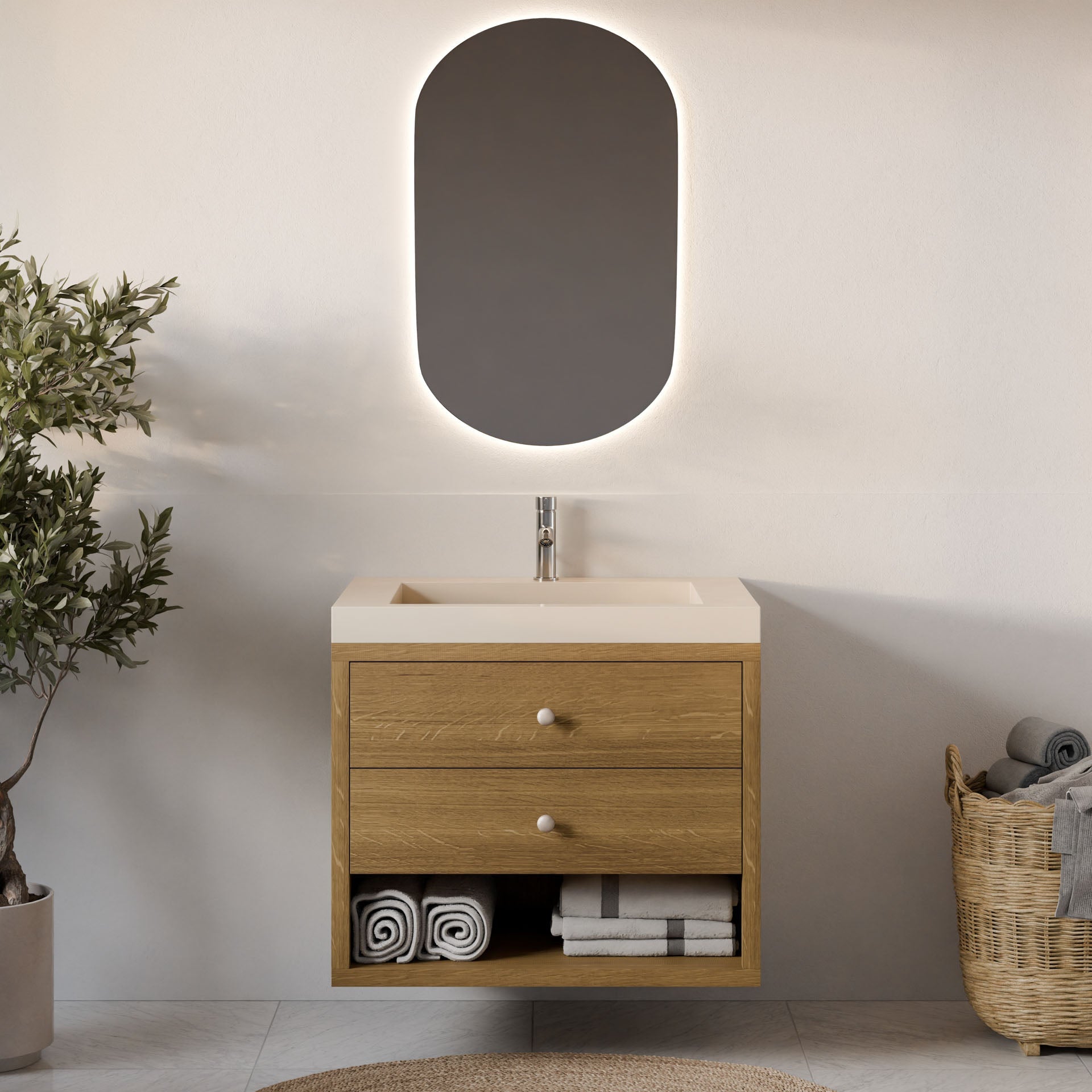 A minimalist bathroom showcases a custom-size floating vanity, the Double Drawer Open Shelf Vanity in White Oak by Krovel Furniture Co. It's adorned with rolled towels and a shelf. Above the sink is an elegant oval backlit mirror. A plant on the left and a wicker basket with towels complete the calming palette.