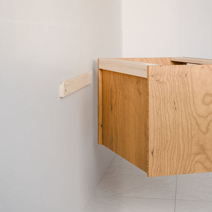 A custom-sized Double Drawer Vanity in Cherry by Krovel Furniture Co., with an open back revealing part of its interior support beam, is being mounted on a white wall. The hardwood veneer gives it a floating appearance, while the floor beneath has a light, speckled pattern.