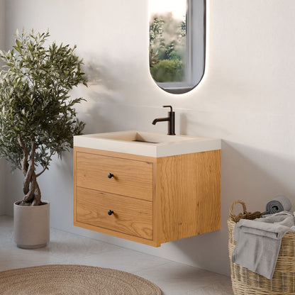 A modern bathroom corner showcases the Krovel Furniture Co. Double Drawer Vanity in Cherry with two custom-size drawers and an oval mirror above. Nearby, a small potted olive tree stands next to a wicker basket with rolled towels on a light-colored floor.