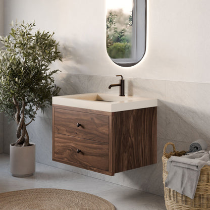 A modern bathroom showcases Krovel Furniture Co.'s Double Drawer Vanity in Walnut with a white sink. A mounted black faucet, a basket of towels, and an olive tree decor adorn the space. An oval mirror reflects natural light, enhancing the custom-sized design.