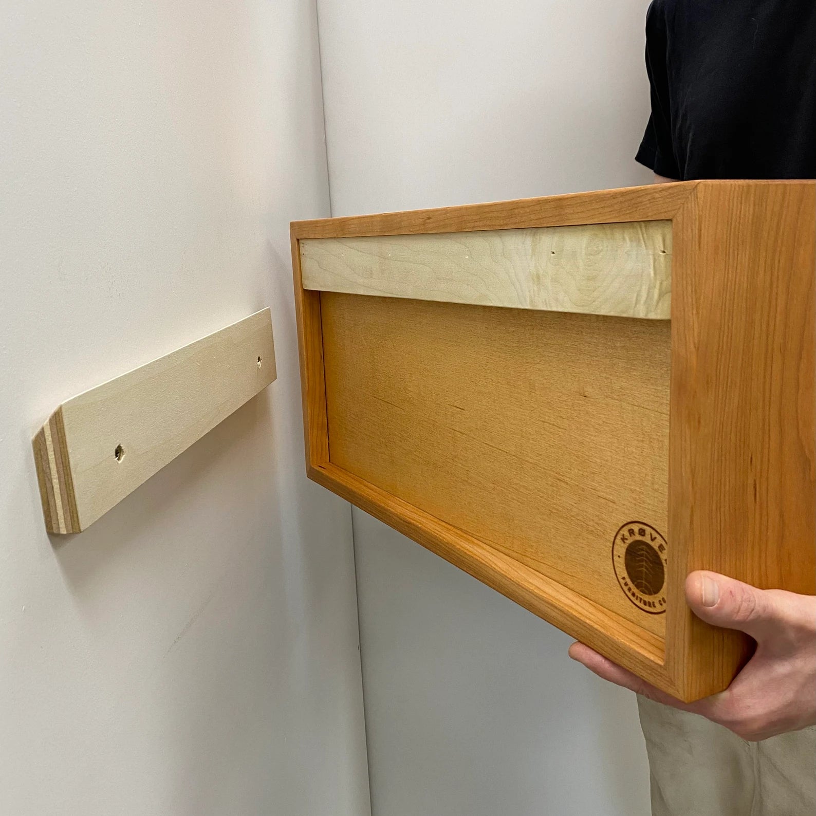 A person holds Krovel's Entryway Organizer in Cherry, a wooden box-like shelf featuring a groove for French-Cleat mounting. A matching mount is attached to the wall nearby, elegantly displaying this floating piece with a circular logo on one side.