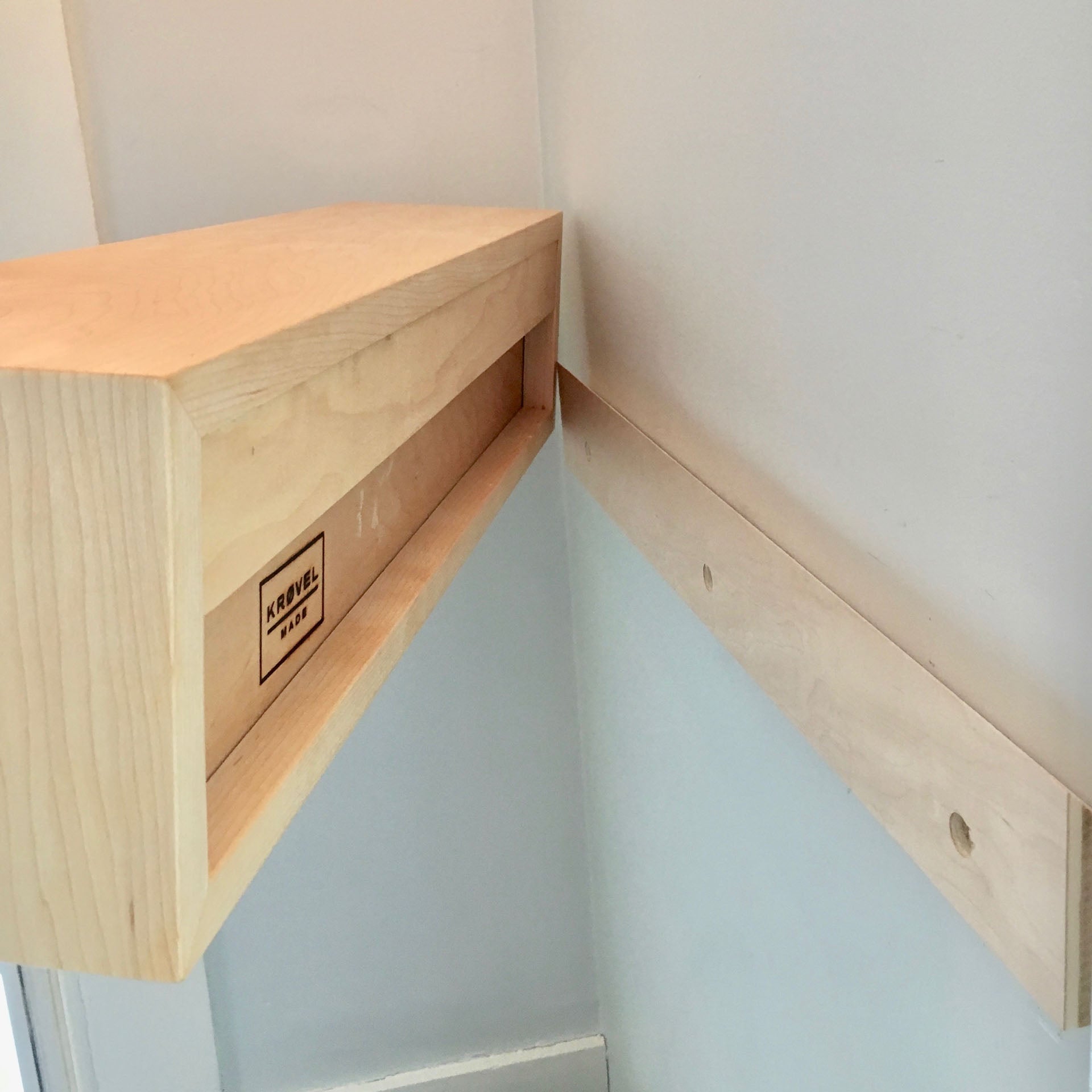 A partially installed floating corner shelf made from American White Oak is on a white wall, featuring a logo with "Kane 1920." The mounting bracket, similar to a French-cleat, has pre-drilled holes.