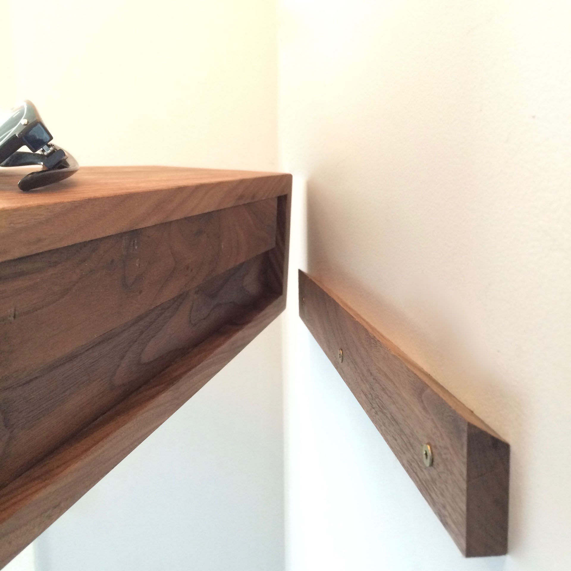 A close-up shows Krovel's Entryway Organizer in Walnut on a white wall. This elegant floating shelf, aligned with a wooden bracket and mounted using the French-cleat technique, holds a small watch on its left end.