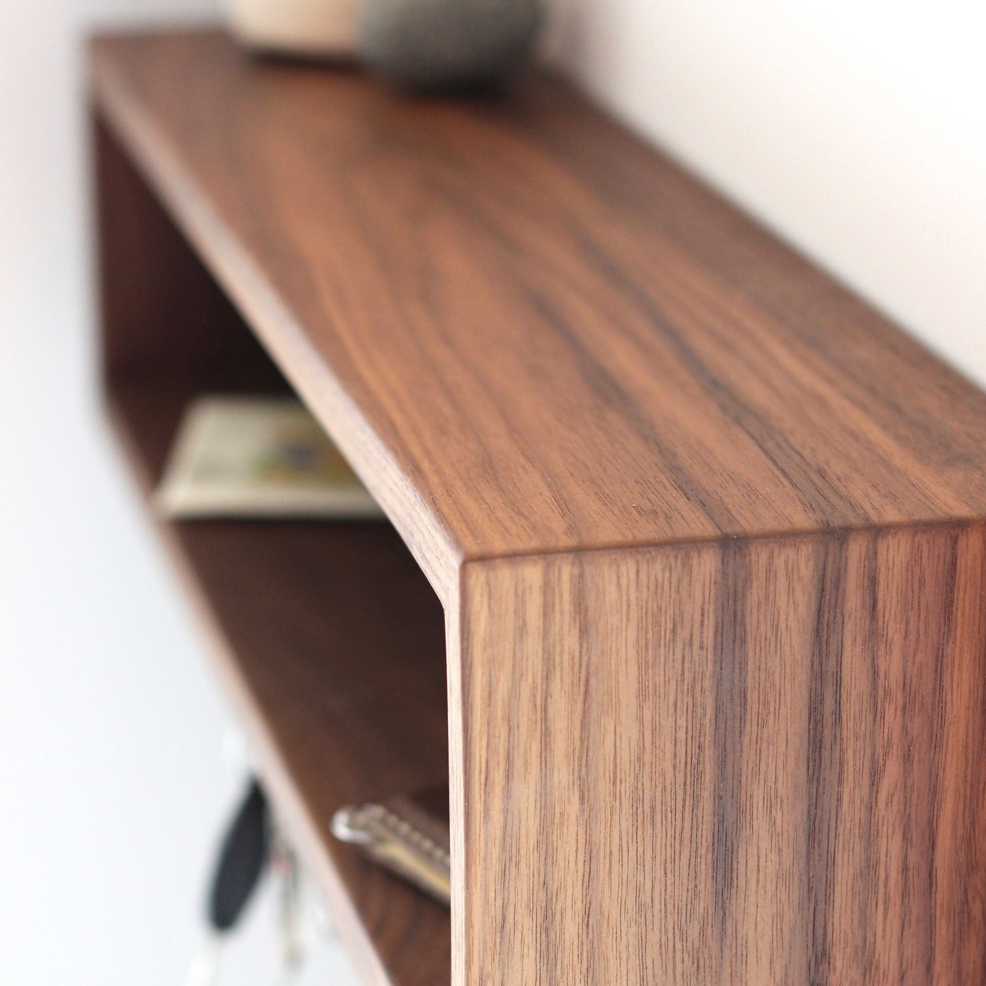 Close-up of the Krovel Entryway Organizer in Walnut, featuring a smooth finish, mounted on a white wall. On this handmade American Black Walnut floating shelf are various small items like a rock and keys, acting as stylish accents. The perspective highlights the rich grain and texture of the wood.