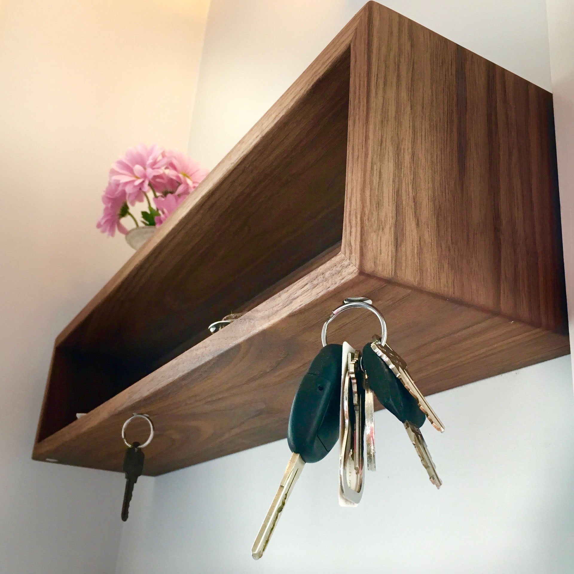 The Krovel Entryway Organizer in Walnut is a wooden wall-mounted shelf crafted from American Black Walnut, featuring hooks for keys underneath. It elegantly holds a small vase with pink flowers, showcasing its sleek design against a plain background.