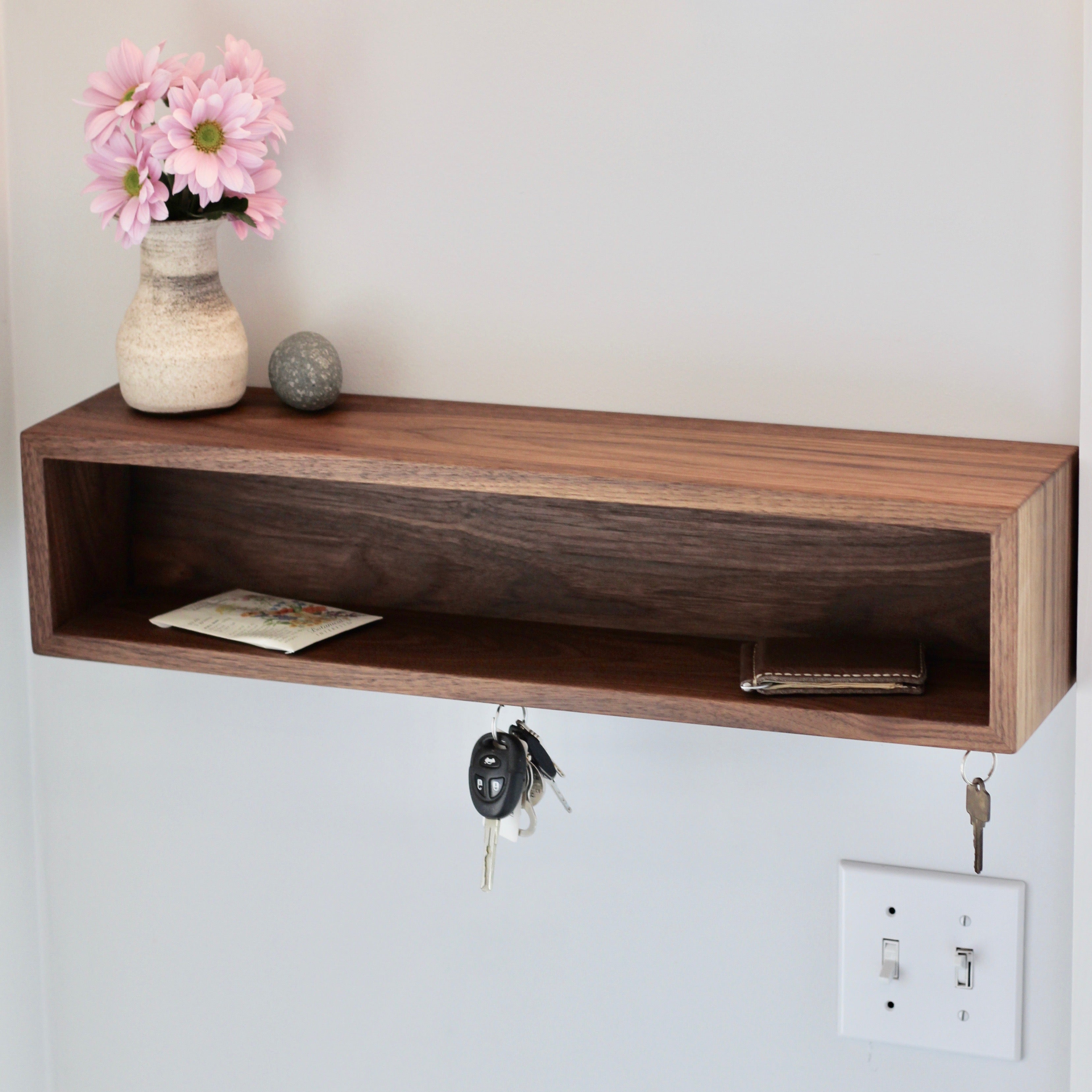 The Krovel Entryway Organizer in Walnut is a minimalist, handmade wall-mounted piece crafted from American Black Walnut. It elegantly holds a small vase with pink flowers and a round stone ornament, while also featuring hooks for keys. An envelope and small item rest on the shelf, conveniently located near a light switch.