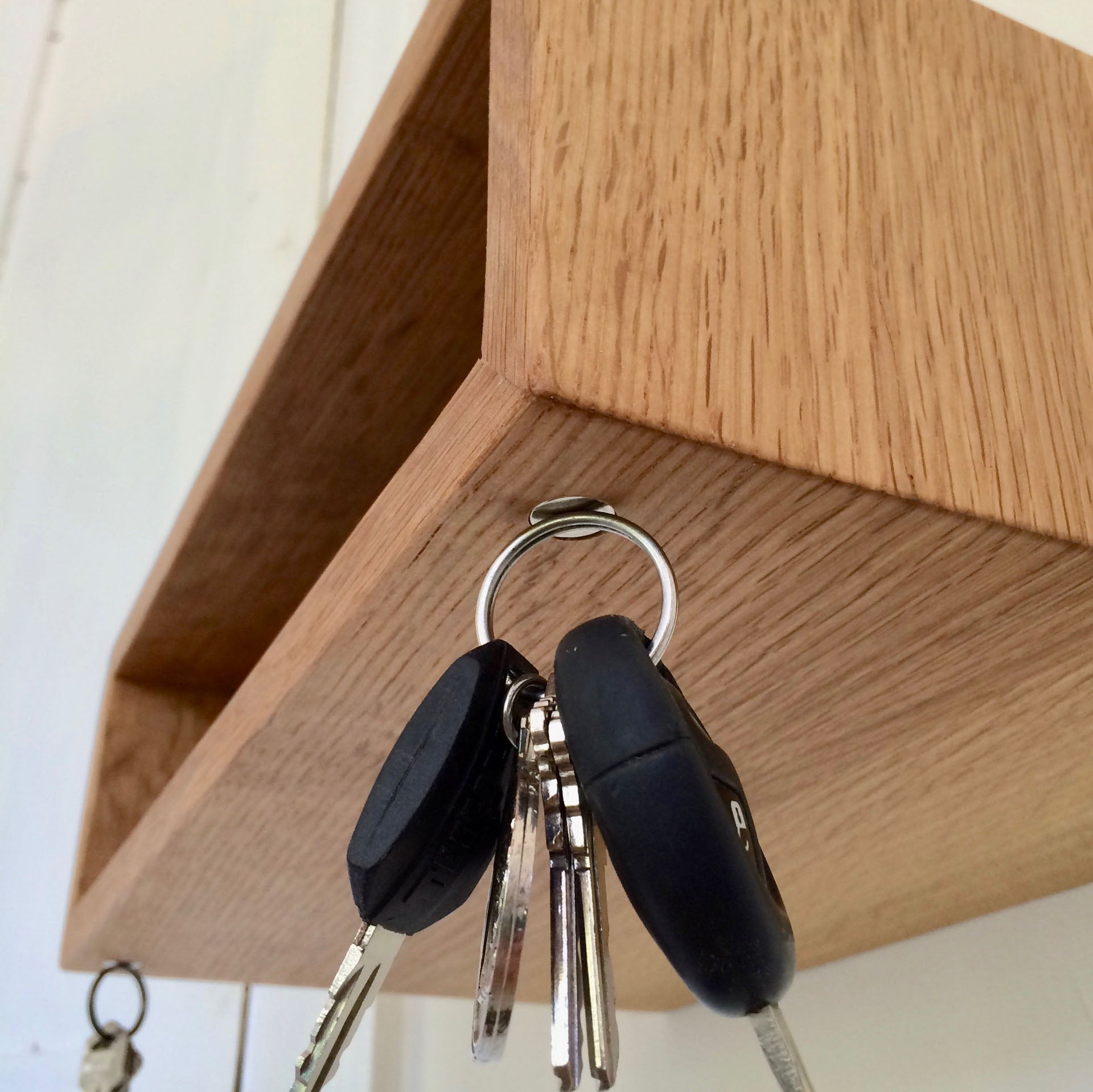 Close-up of a Krovel Entryway Organizer in White Oak, featuring a handmade wooden shelf corner. Car keys, complete with black electronic fobs and multiple metallic key blades attached to a silver ring, hang on a hook beneath the shelf, making it an ideal floating entryway solution.