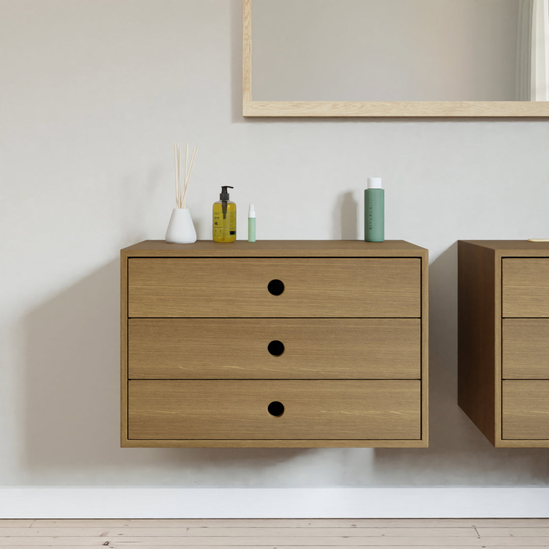 The White Oak Floating Dresser by Krovel Furniture Co. features three spacious drawers and is beautifully paired with a mirror above, creating a sleek wall-mounted look. On top, it elegantly holds various toiletries such as a soap dispenser, lotion bottle, and reed diffuser. Custom sizing is available to achieve the perfect fit for your space.