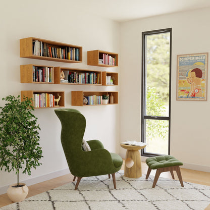 A cozy reading nook showcases a green armchair with an ottoman, accompanied by a small wooden side table and a rug. The walls are adorned with Krovel's Floating Bookshelf in Cherry, filled with books. A large window and a colorful poster enhance the bright, minimalist atmosphere completed with handmade American Cherry accents.