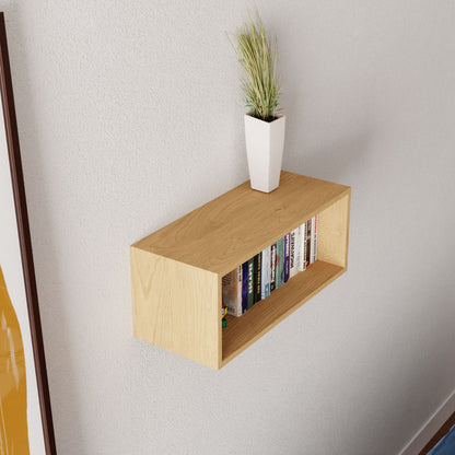 A Krovel Floating Bookshelf in Maple, crafted from American Hard Maple, supports several books and a small white planter containing an artificial grass plant. The background showcases a light-colored wall and the edge of a framed picture, contributing to the minimalist design that gives the impression of sleek floating bookshelves.