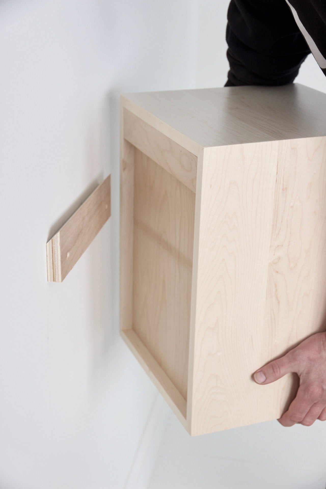 A person is installing a Krovel Floating Bookshelf in Maple on a white wall using a French cleat. The hand is visible aligning the shelves with the cleat already secured to the wall.