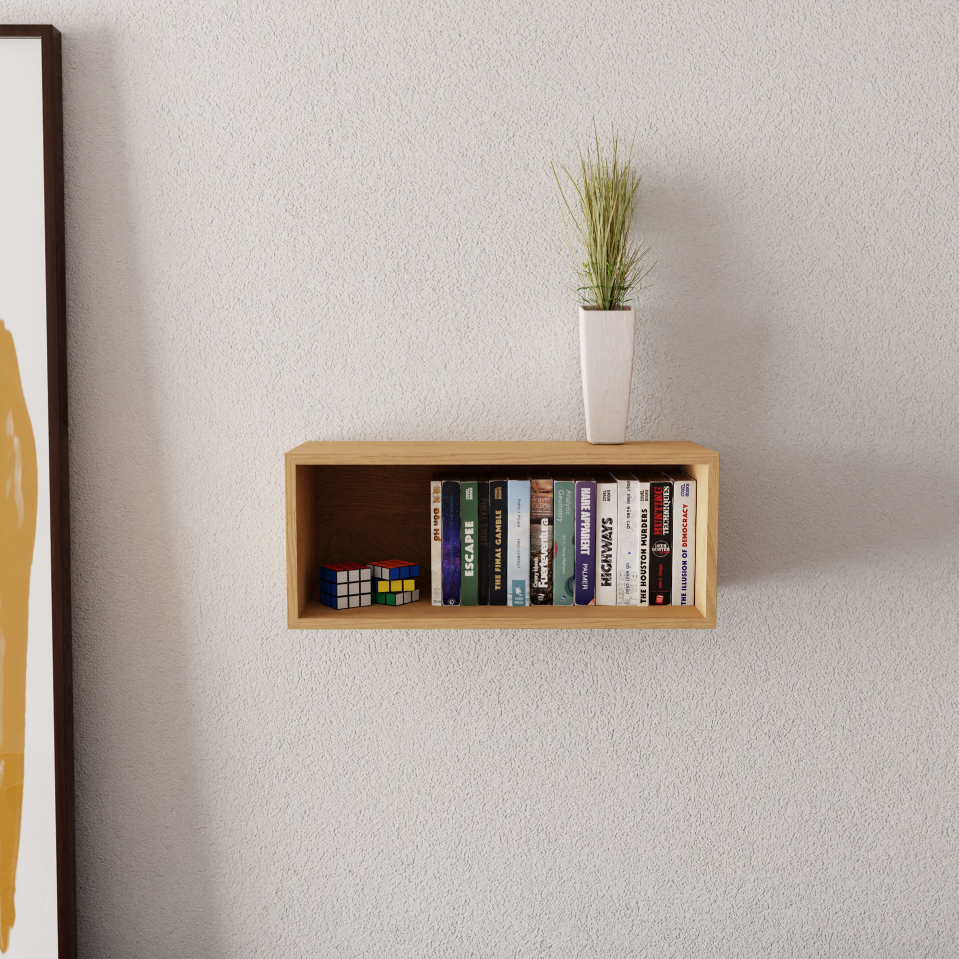 A wall-mounted "Floating Bookshelf in Maple" by Krovel holds books, a tall-grass plant, and a Rubik's cube. The textured gray wall features part of framed artwork on the left.