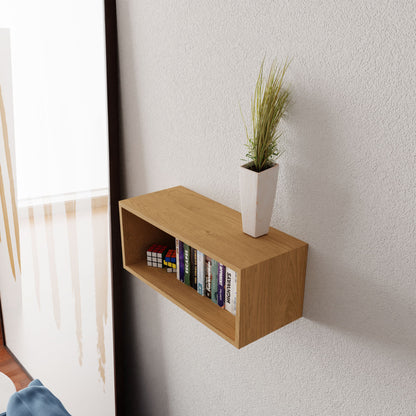 The Krovel Floating Bookshelf in Maple elegantly displays a small potted plant and a collection of books, accompanied by a Rubik's Cube and framed picture. This wooden wall-mounted shelf, crafted from American Hard Maple, is securely attached to the textured white wall using a French cleat.