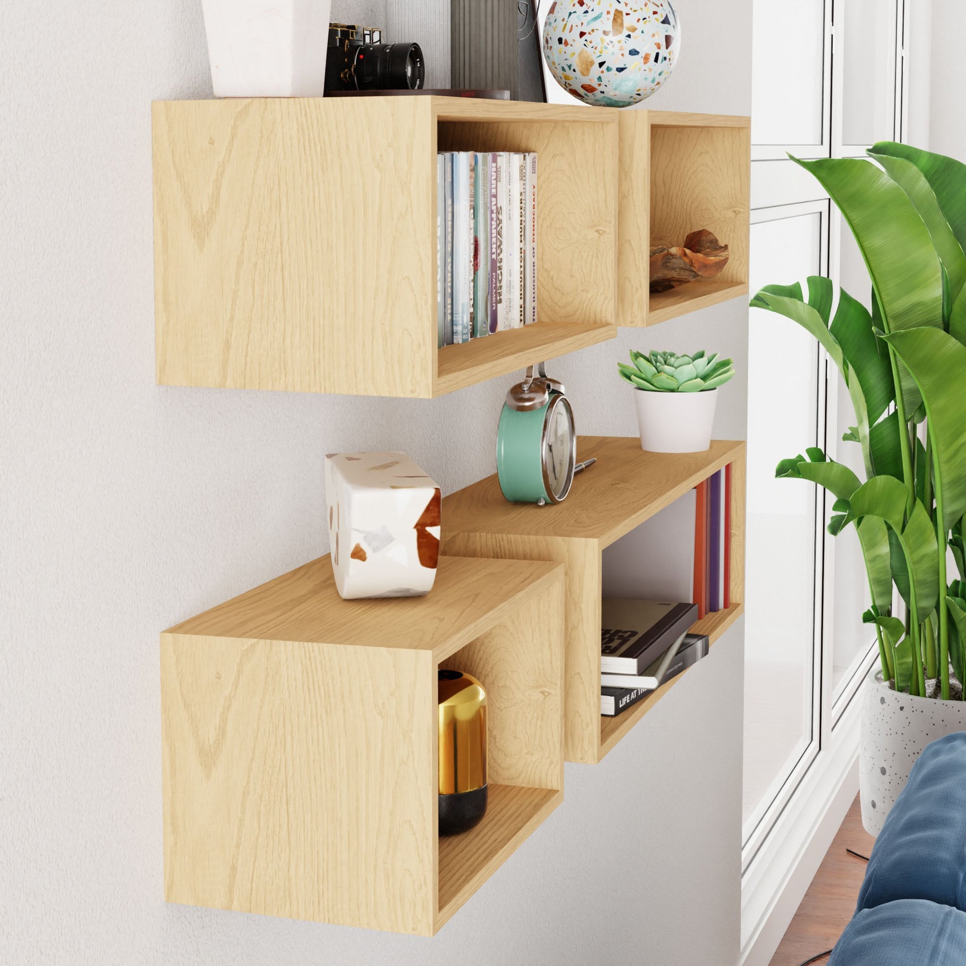 A Krovel Floating Bookshelf in Maple is elegantly mounted on the wall, featuring staggered compartments filled with books, an alarm clock, decorative items, plants, and a globe. Close by, a green potted plant sits beside a window bathed in natural light.