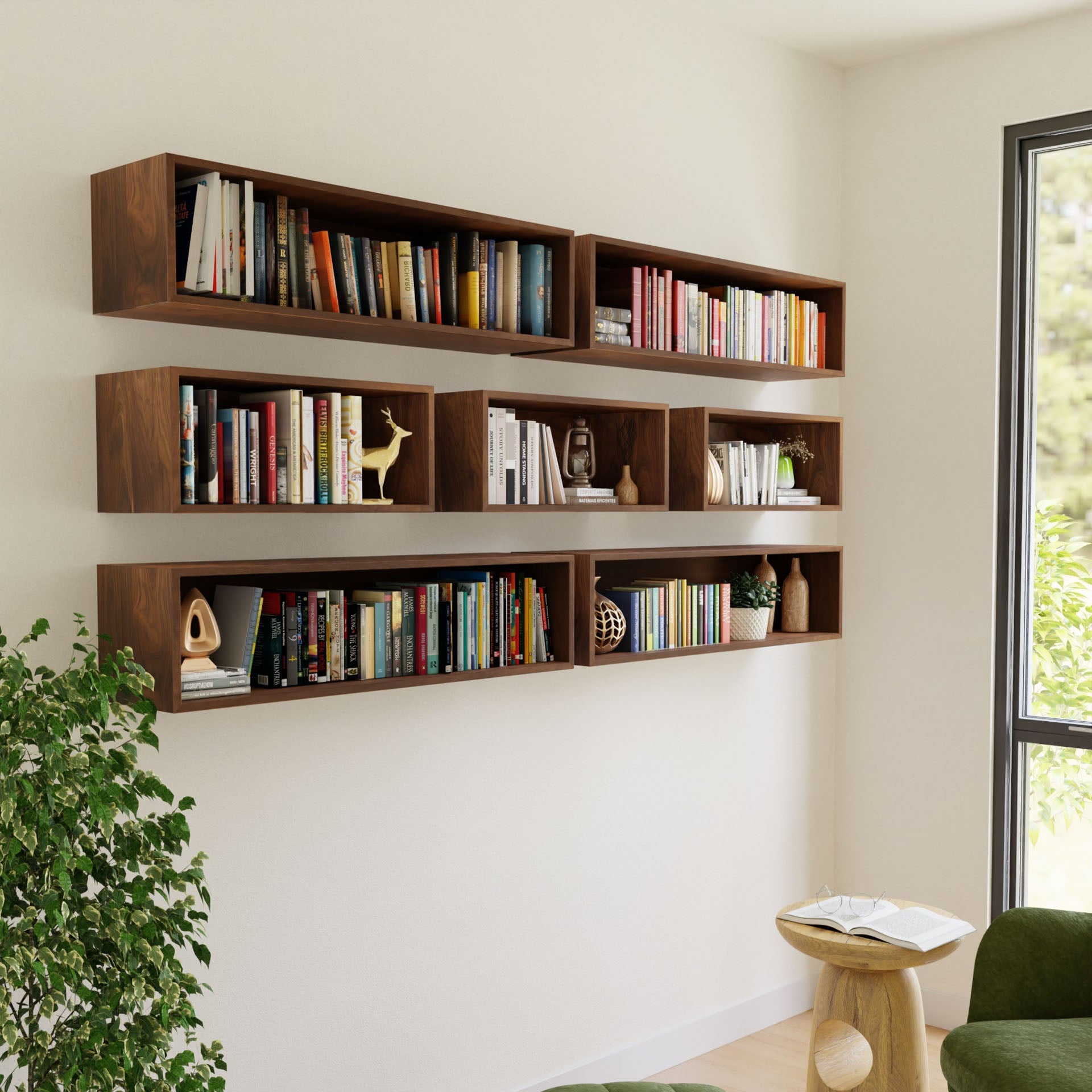 The Krovel Floating Bookshelf in Walnut, crafted from American Black Walnut, displays books and decor items, such as small sculptures and plants on three staggered rows. Nearby, a green chair and a small side table with a book complement the setup while a large window offers lush greenery views.