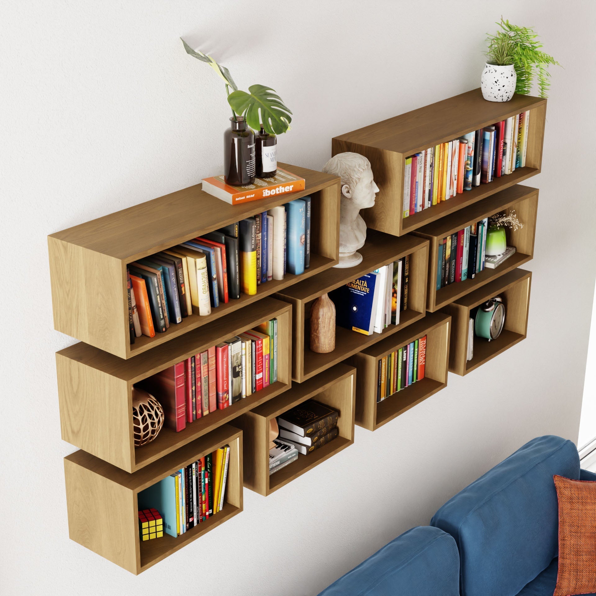 The Krovel Floating Bookshelf in White Oak is a modern, wall-mounted design with staggered compartments filled with books. Handmade, it features a small plant, decorative bust, and candle on top. A glimpse of a blue sofa enhances its stylish look.