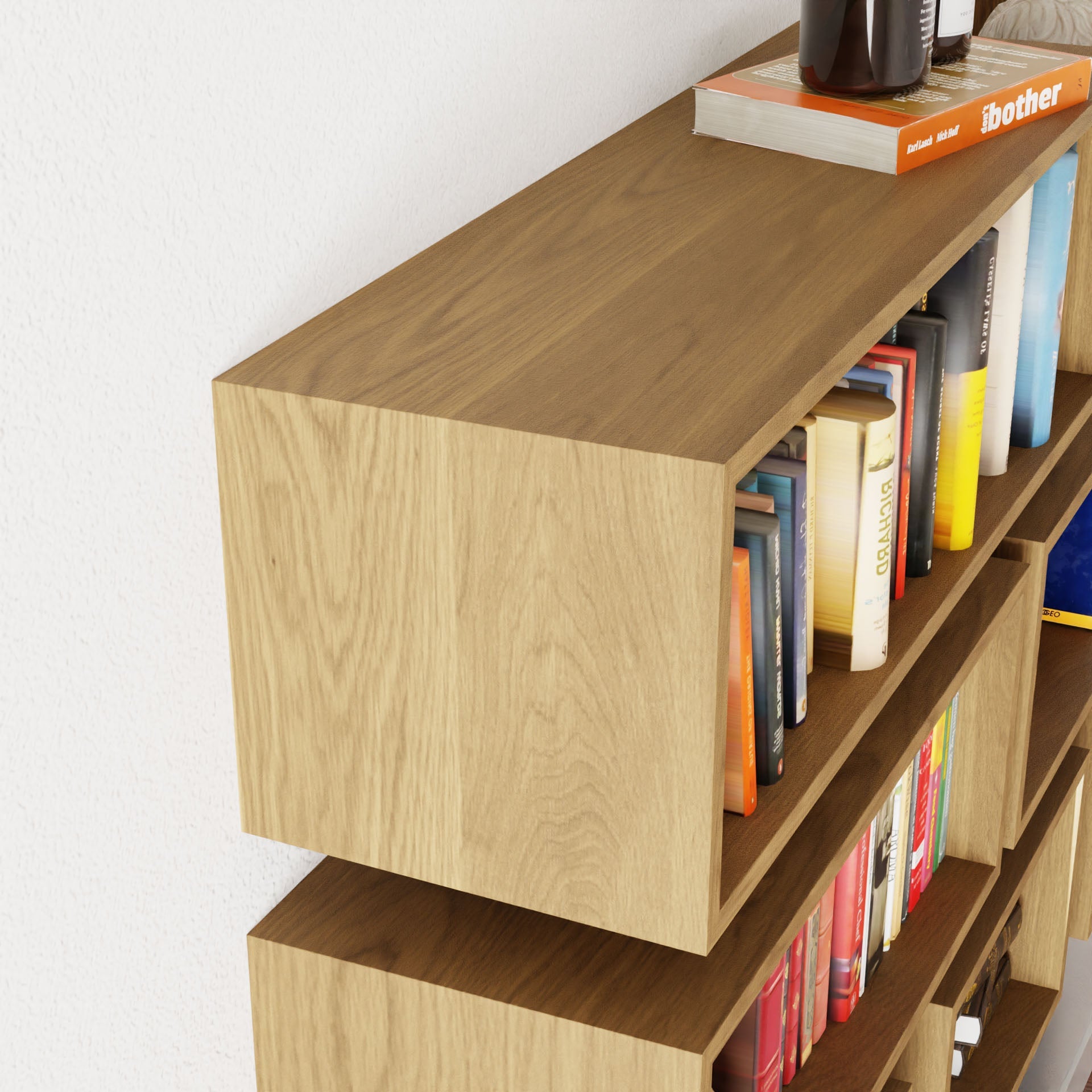 The Krovel Floating Bookshelf in White Oak, minimalist and handmade, is filled with books. Magazines and a black bottle rest on top, while the bookshelf leans gracefully against a white wall, showcasing neatly arranged upright books.