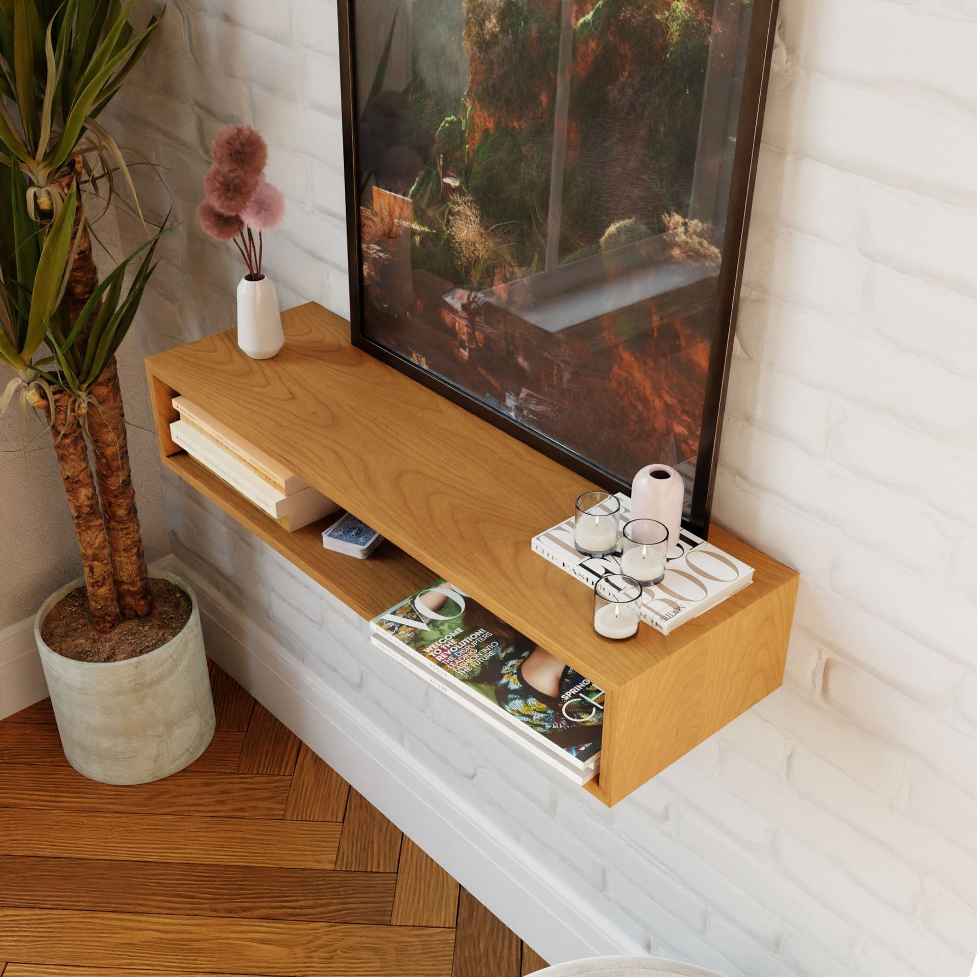 A chic Floating Console Table in Cherry by Krovel, inspired by mid-century modern design, displays decor items such as candles, a small white vase, and books against a white brick wall. A potted plant with tall green leaves is placed beside the table. Natural light accentuates the warm tones of American Cherry wood and flooring.