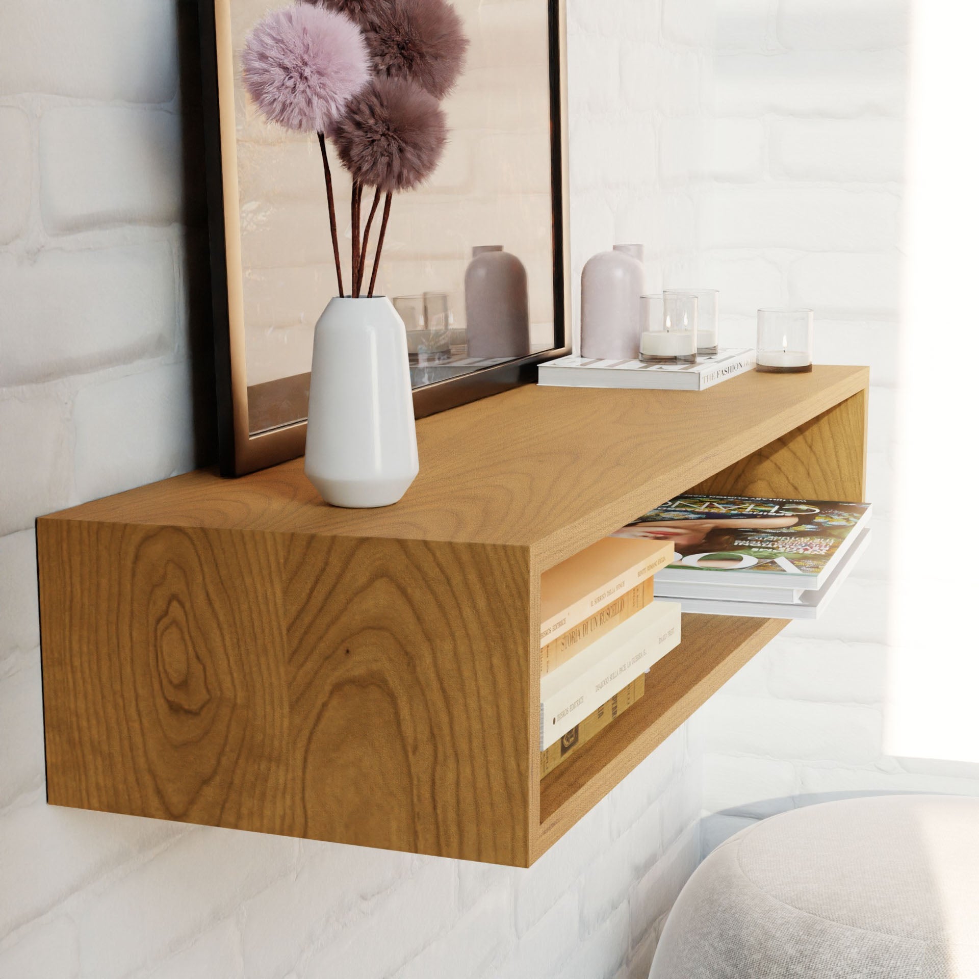 A Krovel Floating Console Table in Cherry on a white brick wall displays a white vase with pink flowers, a candle, decor items, books, and magazines. A framed picture is elegantly propped behind the display.