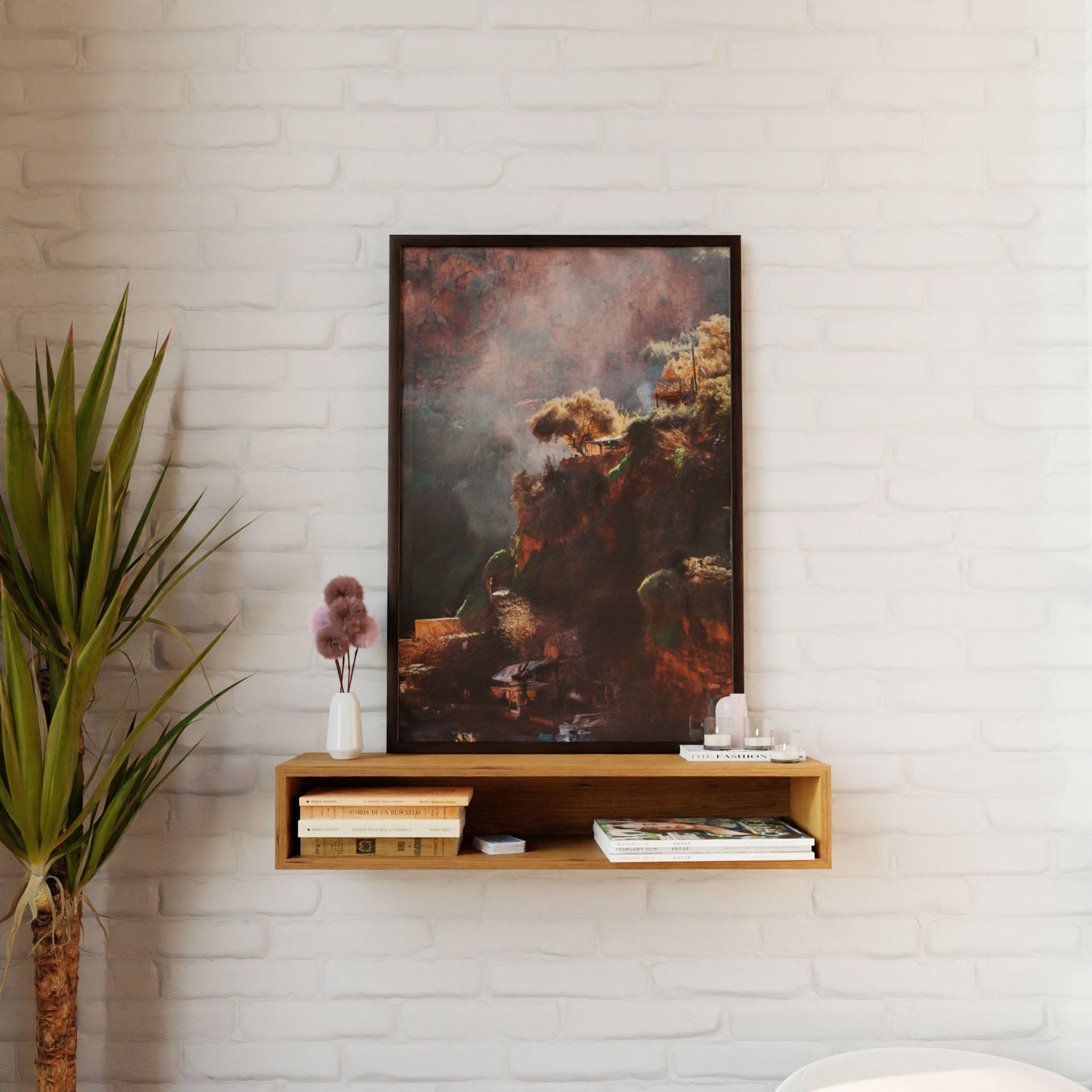 A framed nature painting hangs on a white brick wall. Below, a Krovel Floating Console Table in Cherry holds books, magazines, a small white vase with dried flowers, and a white candle while a tall green plant stands to the left.