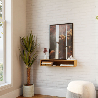 A minimalist corner showcases a tall potted plant next to a Krovel Floating Console Table in Cherry, elegantly displaying decor items and framed artwork on a white brick wall. Natural light streams through the nearby window, creating a serene and airy ambiance.