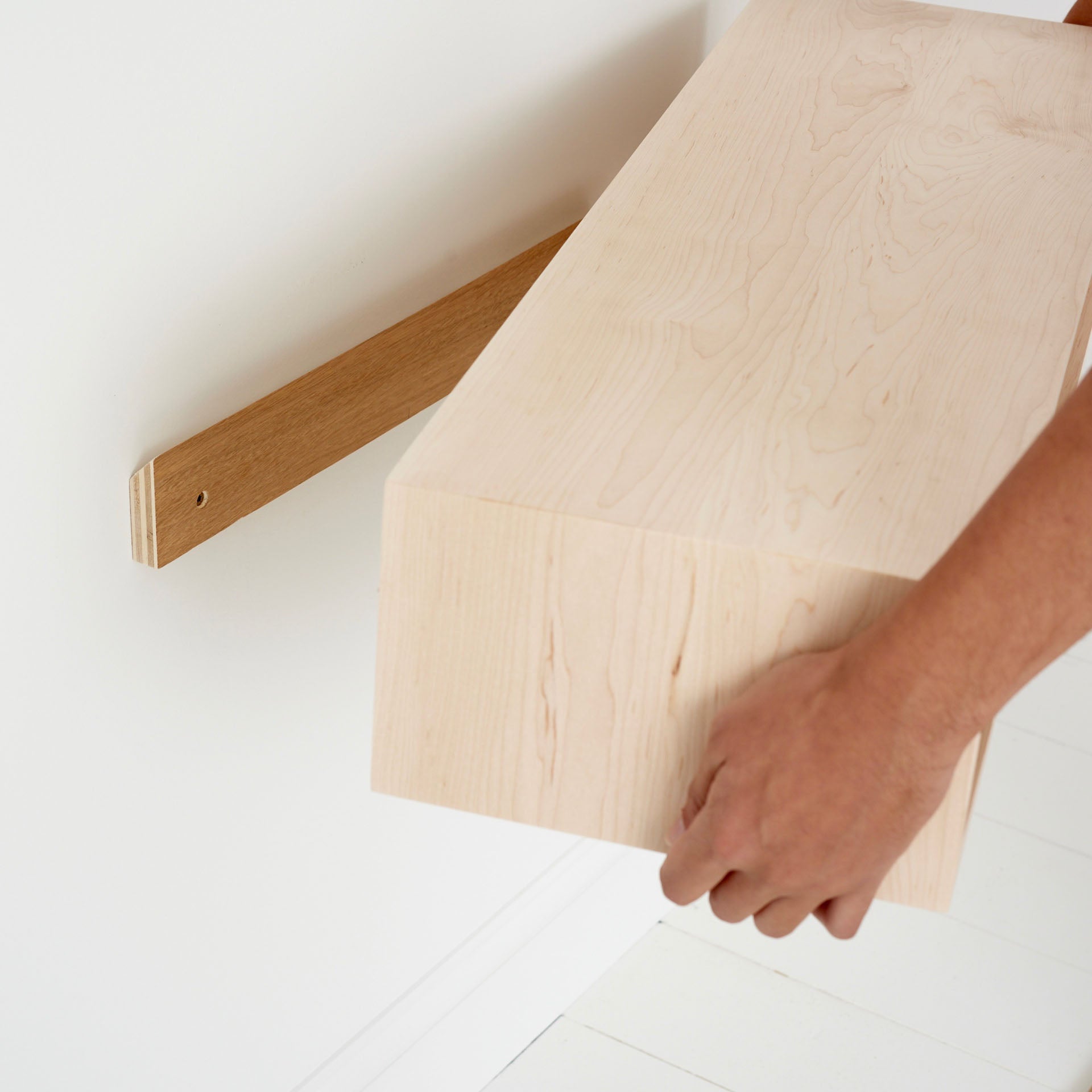 A person installs the Krovel Floating Console Table in American Hard Maple onto a wall bracket. The table's minimalist rectangular design, akin to mid-century modern style, beautifully contrasts with its darker bracket against a white background.