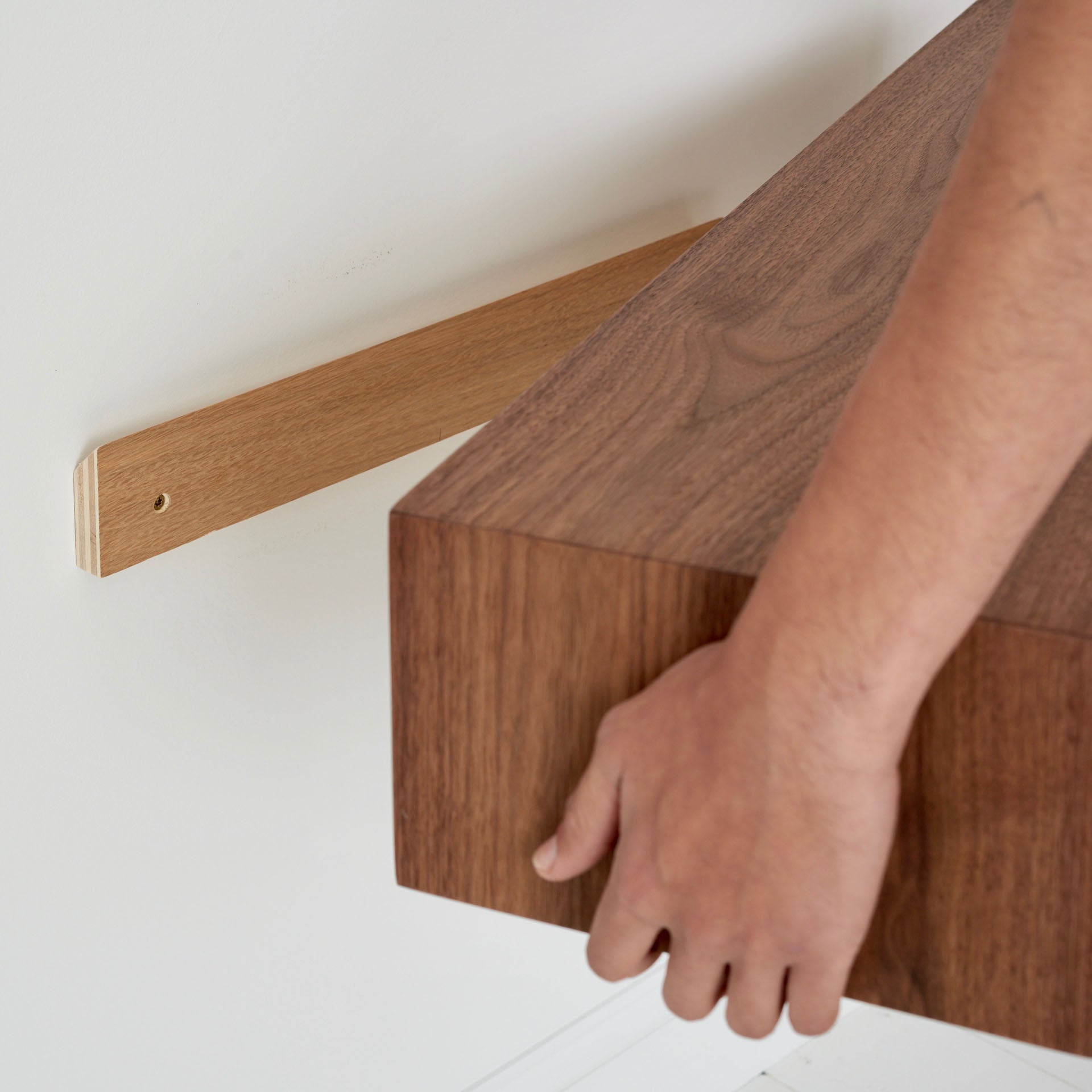 A hand places the Krovel Floating Console Table in Walnut on a mounted bracket against a white wall. The table, made from American Black Walnut, boasts a smooth brown finish and is carefully aligned for installation.