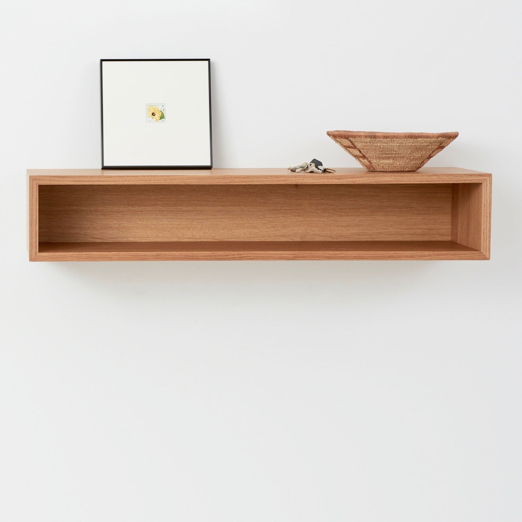 A minimalist wooden console table by Krovel, crafted from elegant White Oak, is mounted on a white wall. It holds a small framed picture, a few objects, and a woven basket. The console table's simple design seamlessly blends with the clean wall, embodying a touch of mid-century modern elegance.