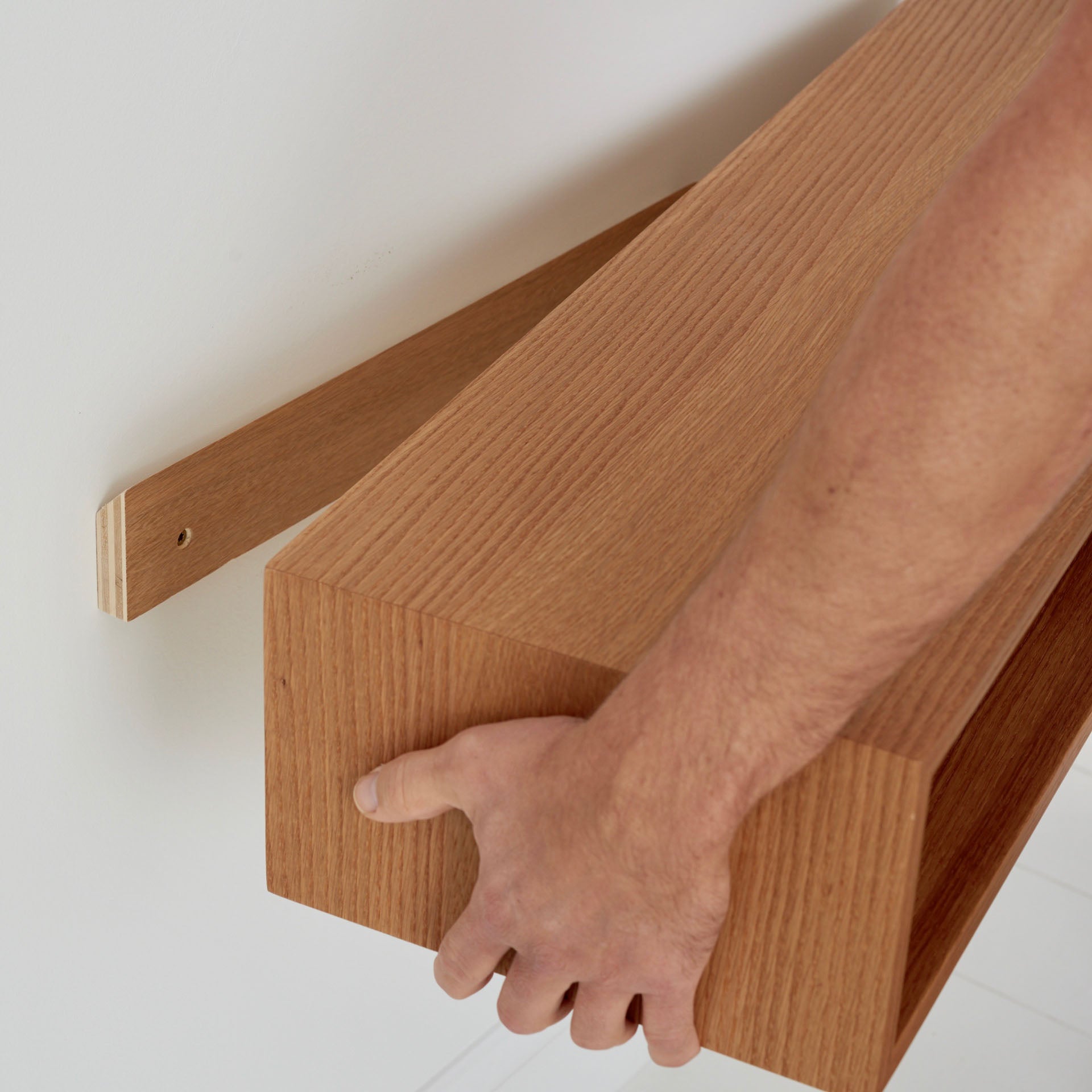 A person is installing a wooden floating console table from Krovel on a white wall. Crafted from White Oak, the hollow design allows it to fit seamlessly over a hidden bracket. With one hand, the person skillfully positions and adjusts the console table for proper alignment, ensuring a perfect fit.