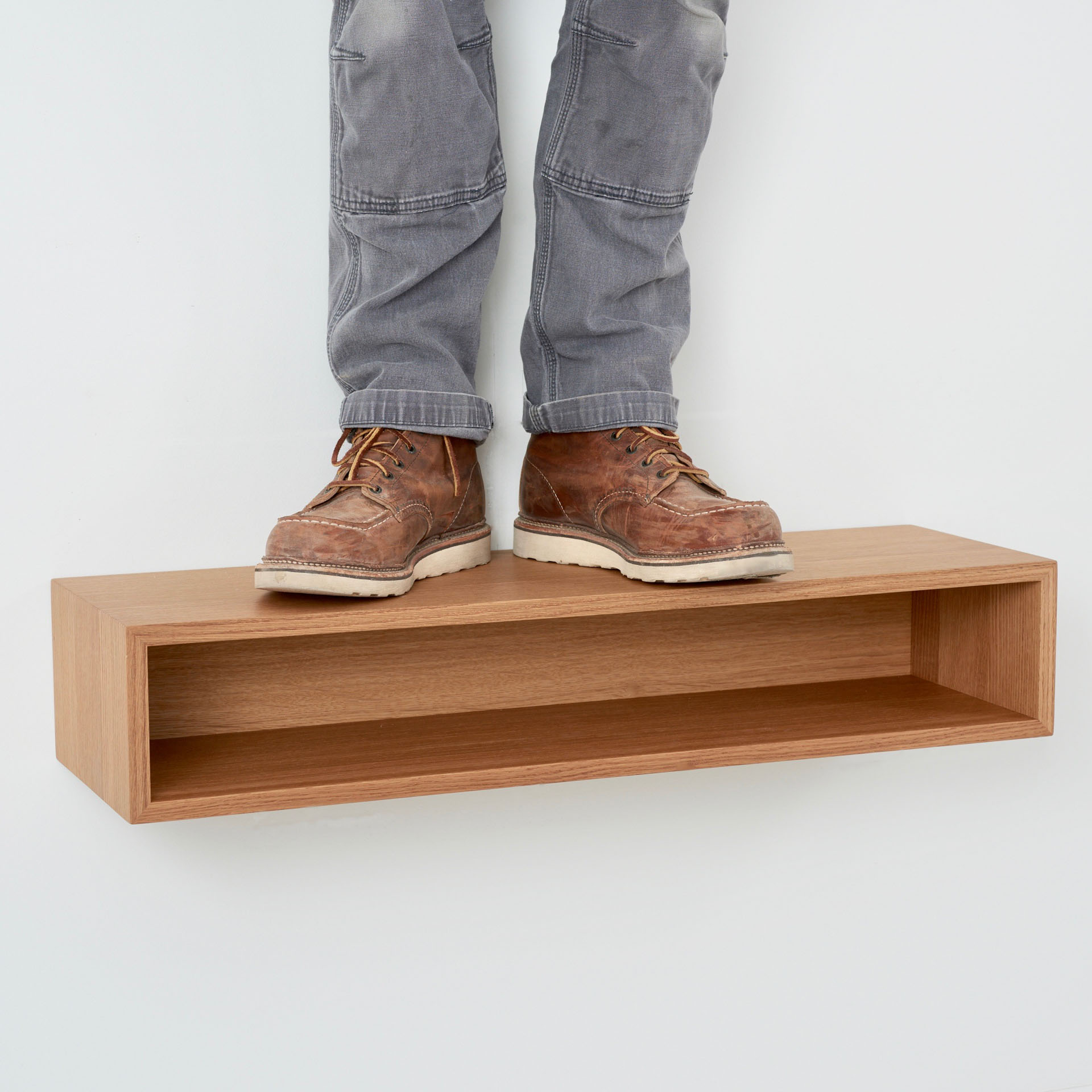 A person in jeans and brown boots stands on a wooden shelf, resembling a white oak Floating Console Table by Krovel, mounted on a light-colored wall.