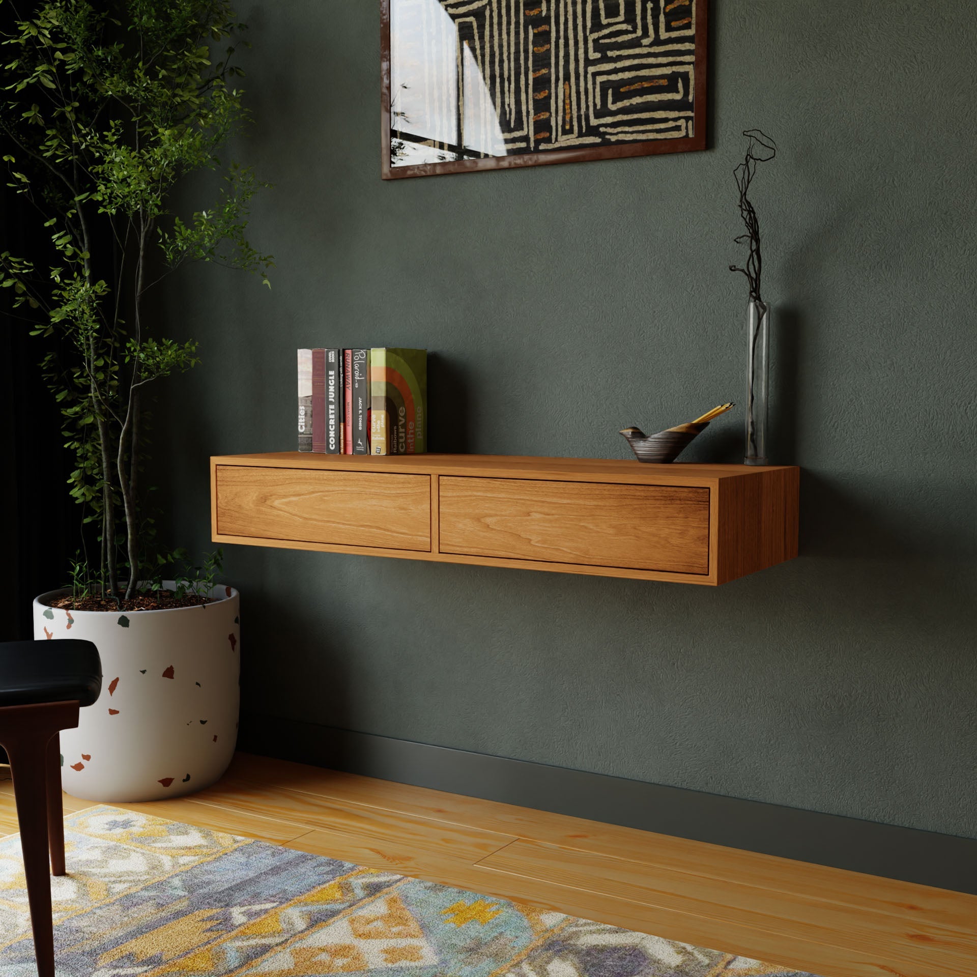 A minimalist room showcases a Krovel Floating Desk in Cherry, featuring two drawers that perfectly complement an American Cherry shelf adorned with books and a decorative piece. Above, a geometric art piece is displayed. Nearby, a tall plant in a white pot stands on the wooden floor with a patterned rug.