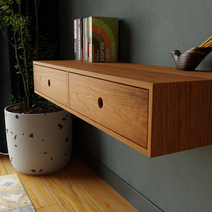 A Krovel Floating Desk in Cherry, featuring two drawers and expertly mounted on the wall, holds a selection of books and a bowl of pencils. Crafted from American Cherry, this handmade wooden piece complements the space beautifully. Below it rests a large potted plant on the wooden floor, introducing greenery to the area.