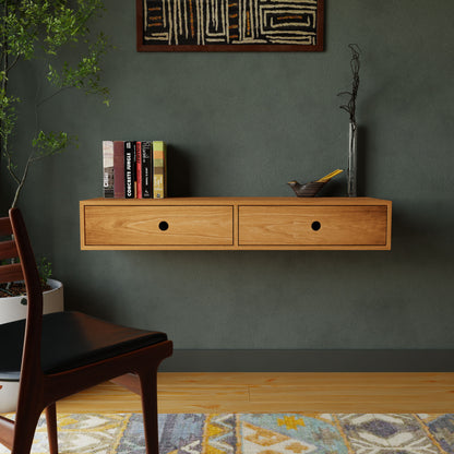 A minimalist room showcases a Krovel Floating Desk in American Cherry, adorned with books and a decorative vase holding a branch. In the foreground, a handmade wooden chair with a black seat is positioned next to a potted plant. The green wall enhances the vibrant appearance of the colorful rug on the wooden floor.