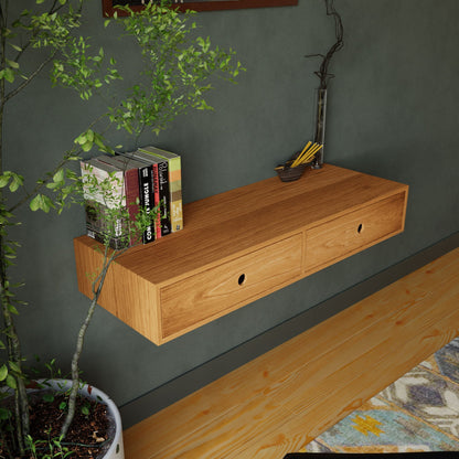 A Krovel Floating Desk in Cherry, featuring two drawers, is mounted on a green wall. The handmade desk holds a few books and a vase filled with pencils. Nearby, a potted plant adds greenery to the cozy room, which is complemented by a light wooden floor and a patterned rug.