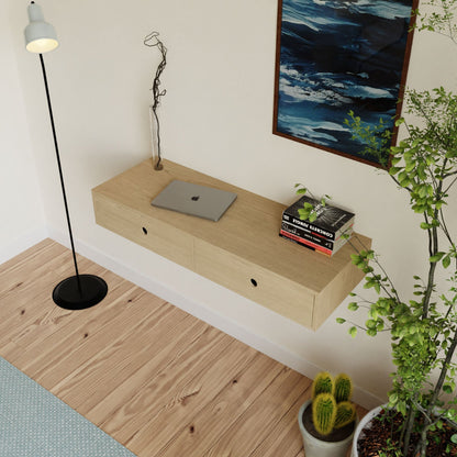 A minimalist room features a light wooden floating desk from Krovel's collection, crafted from Maple and finished with VOC-Free Polymerized Linseed Oil, holding a laptop. A black floor lamp stands nearby with potted plants and cacti enhancing the space, while a framed blue-toned artwork hangs on the wall.