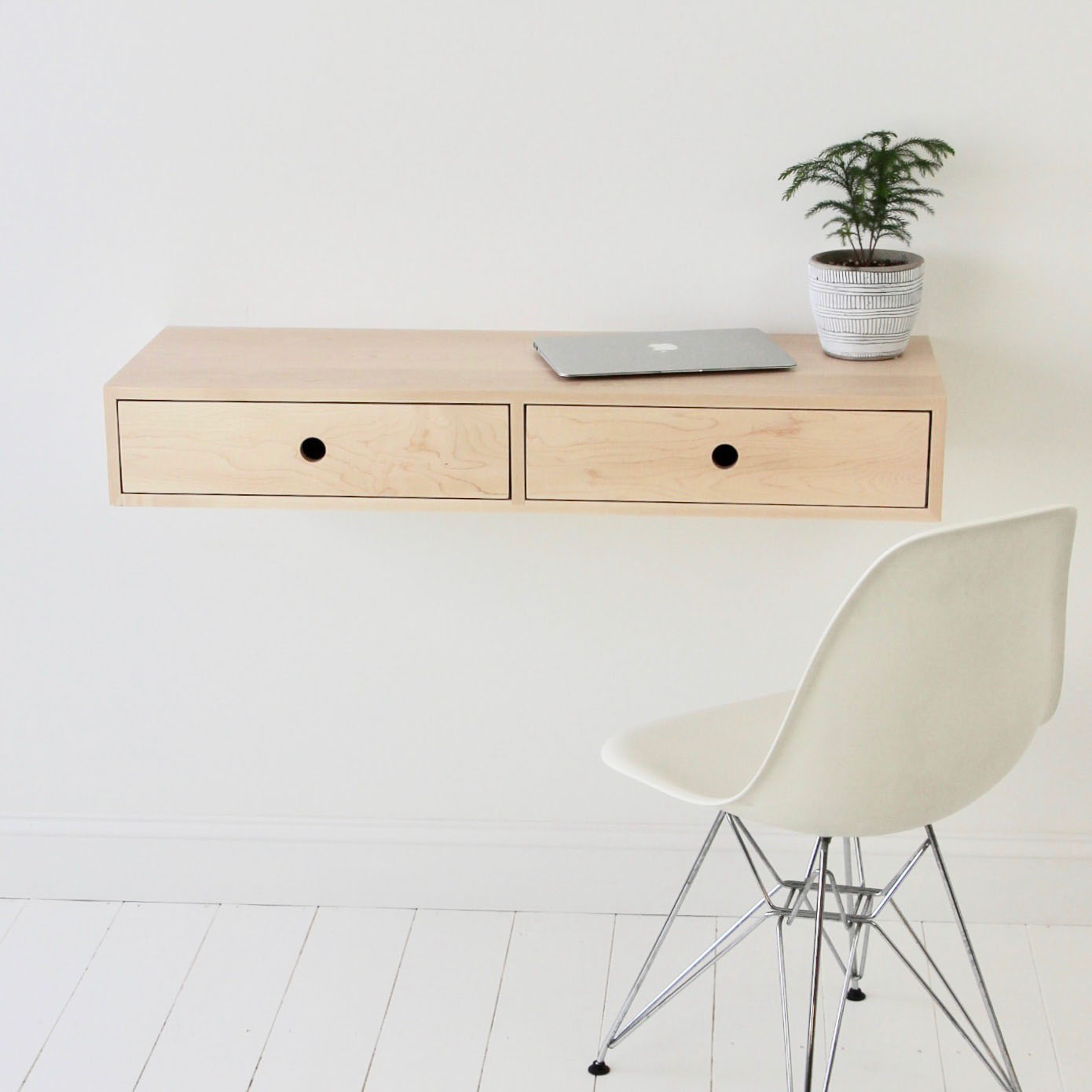 A minimalist workspace features a Krovel Floating Desk in Maple with two drawers. The desk is adorned with a silver laptop and a potted plant, while a white chair with metal legs sits on the pristine wooden floor against a crisp white wall.