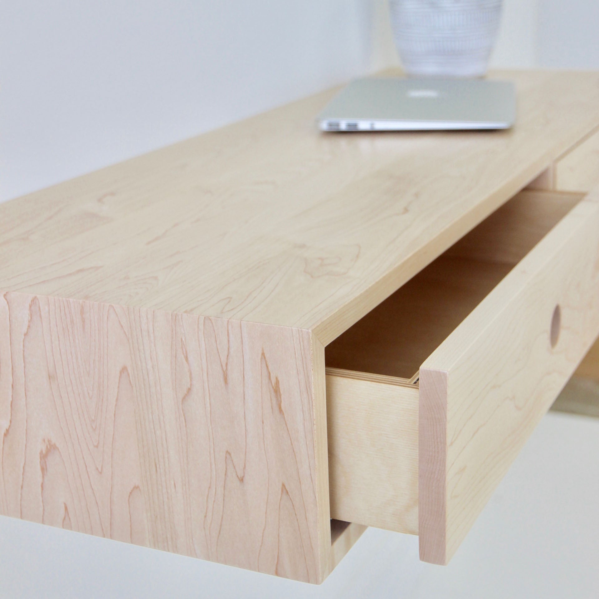 A sleek light wooden floating desk from Krovel, crafted in Maple and finished with VOC-free polymerized linseed oil, showcases an open drawer. Atop it sits a closed silver laptop, while a striped plant pot can be seen peeking from the background. The desk's minimalist design beautifully highlights its craftsmanship.