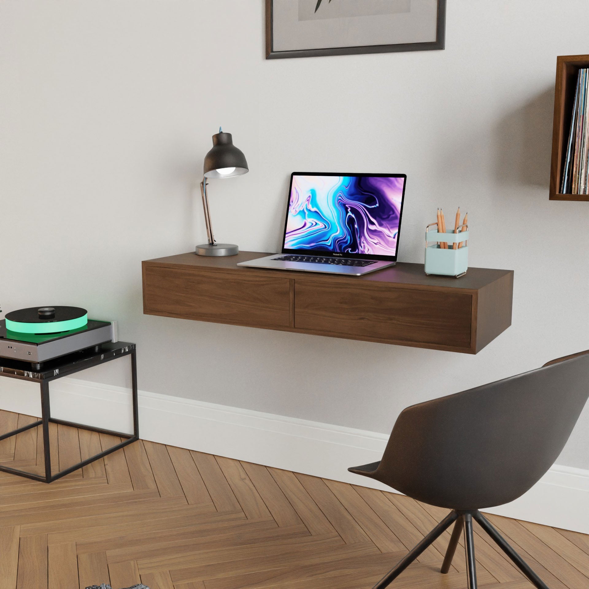 A minimalist workspace features a Floating Desk in Walnut by Krovel, showcasing a laptop with colorful visuals, a desk lamp, and a pencil holder. In front sits a modern black chair, while to the left lies a record player, all elegantly set against the herringbone wood floor.