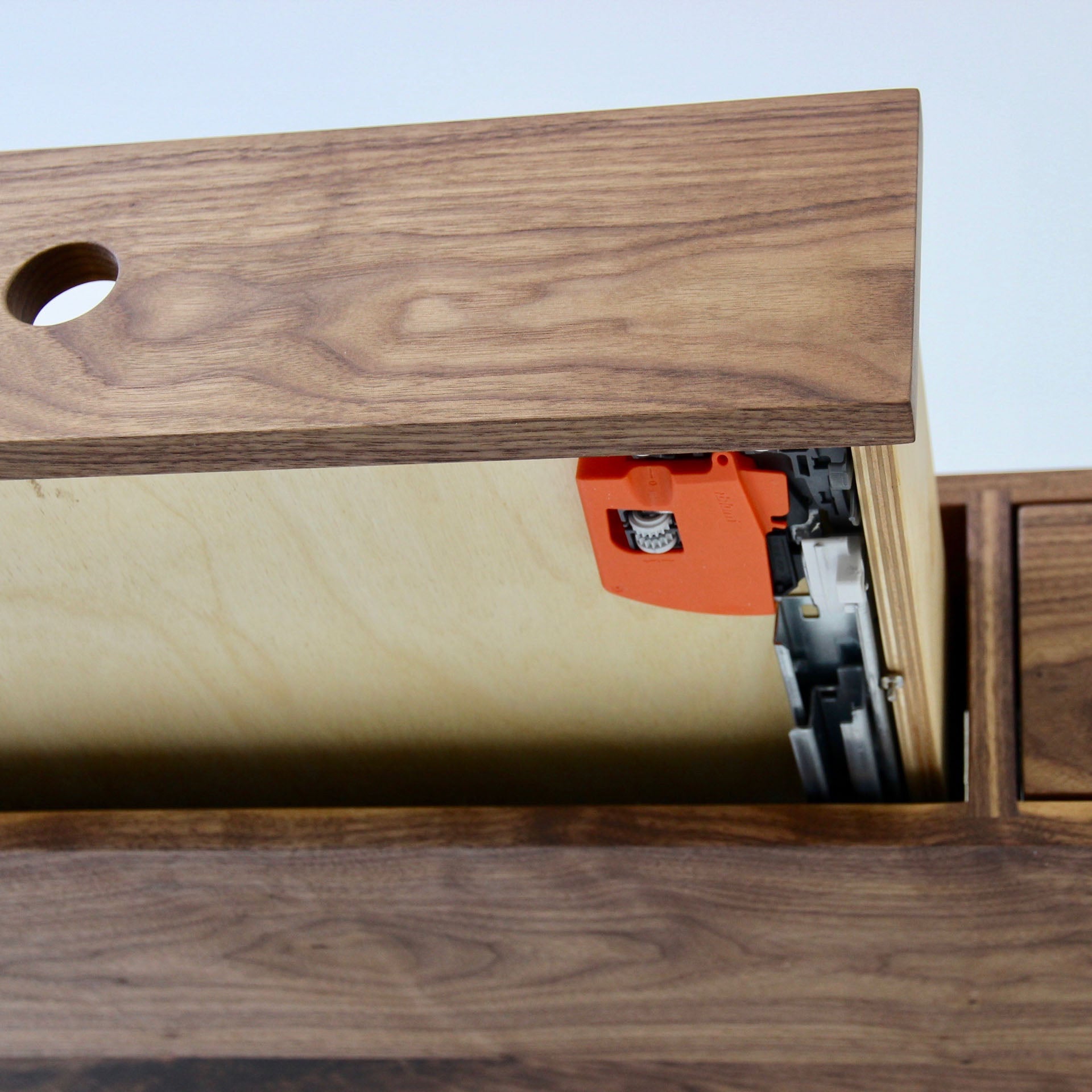 Close-up of a partially open wooden drawer featuring visible dovetail joints and an orange soft-close mechanism, set against the sleek surface of Krovel's handmade Floating Desk in Walnut. The American Black Walnut displays intricate wood grain patterns beneath a circular hole on top.