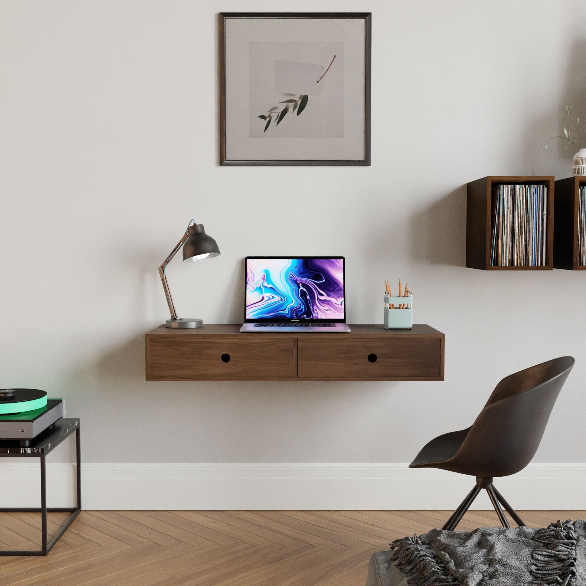 In a contemporary workspace, you'll find the Krovel Floating Desk in Walnut, elegantly holding a laptop with vibrant visuals, accompanied by a desk lamp and a cup filled with pencils. Nearby stands a black chair, while framed art and shelves adorned with vinyl records grace the wall. A record player adds to the ambiance.
