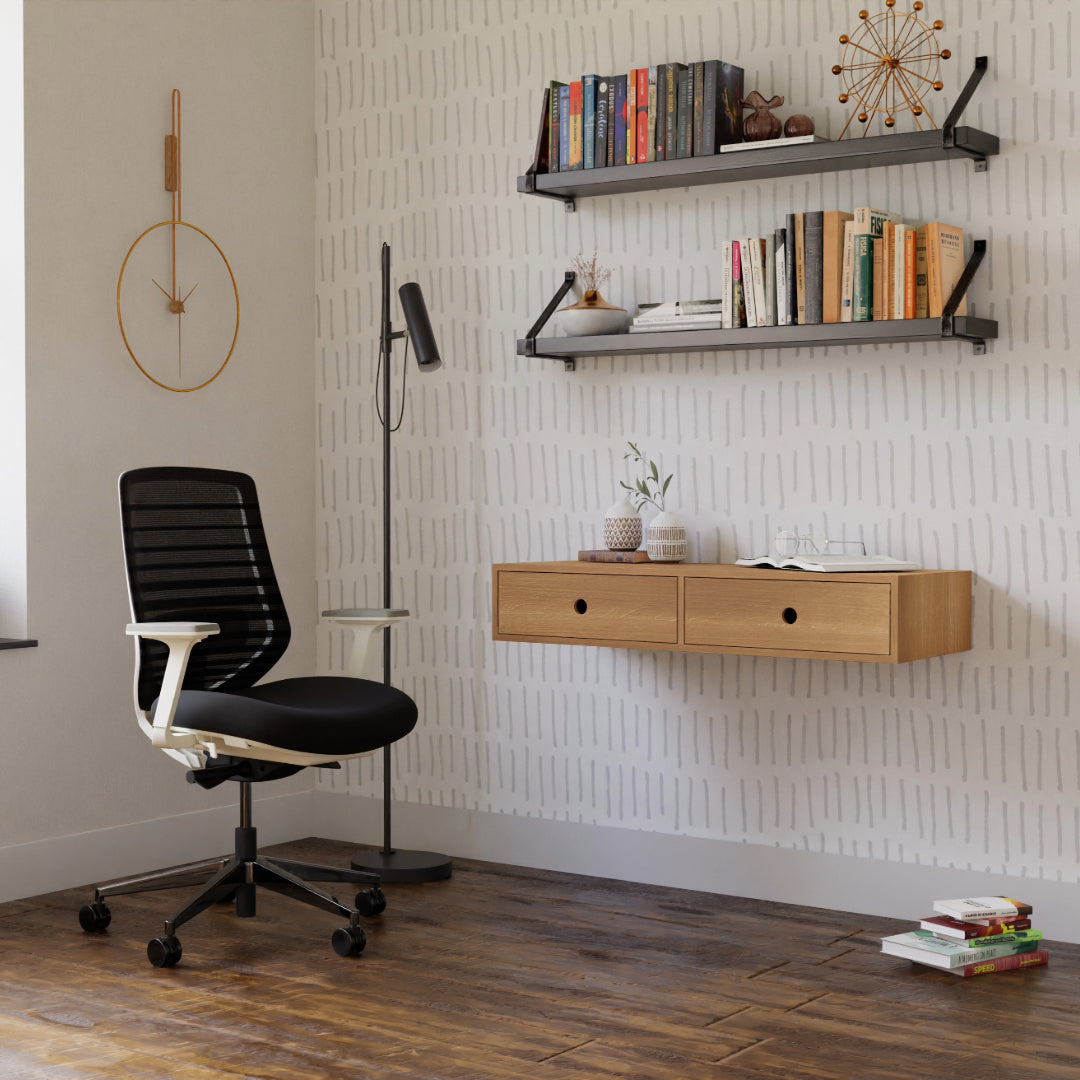 A modern minimalist workspace includes a black office chair, a tall floor lamp, and a Krovel Floating Desk in White Oak with two drawers. Above the desk are two shelves adorned with books and decor. A geometric wall clock hangs against patterned wallpaper, complemented by the warmth of the wooden floor.