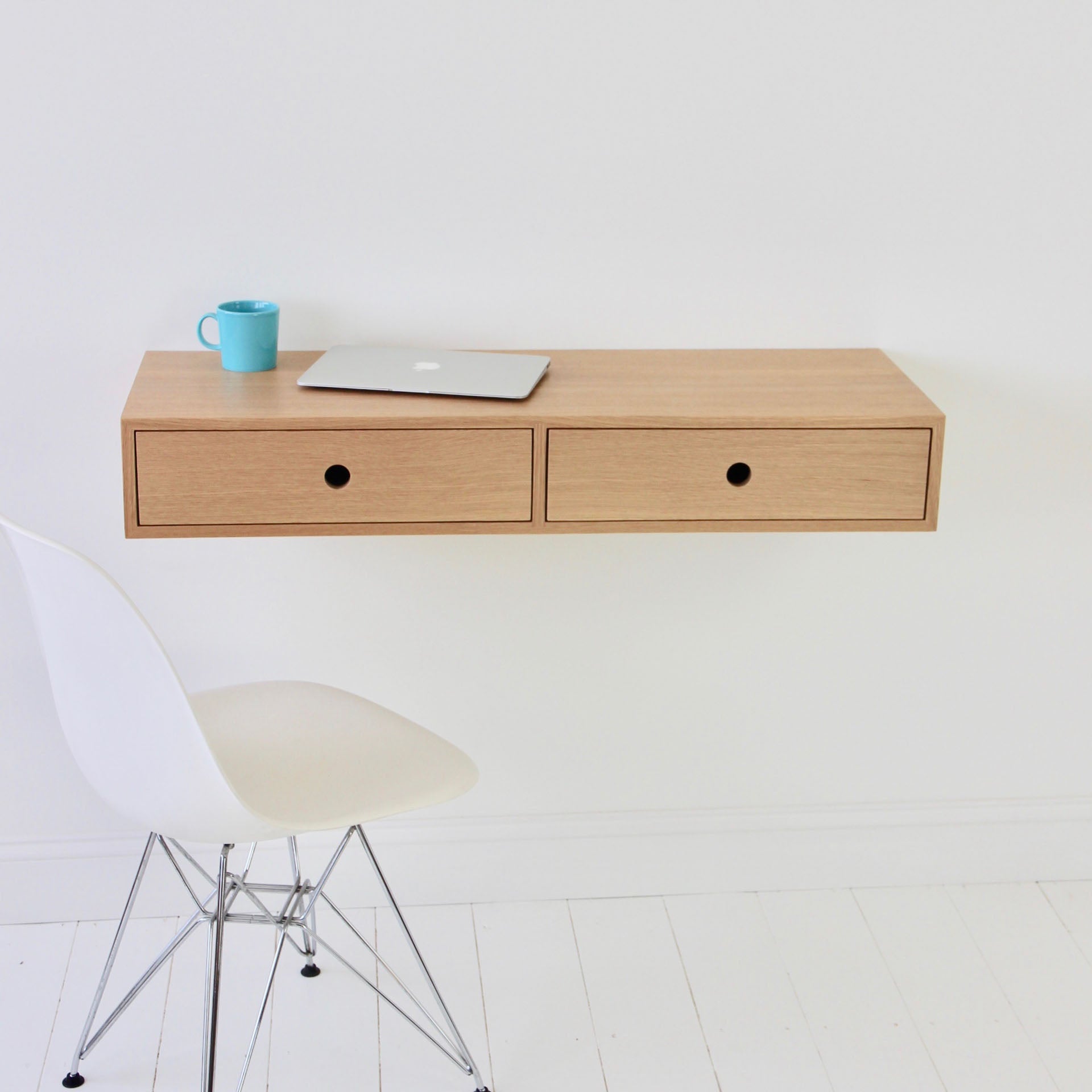 A minimalist workspace showcases a handmade floating desk crafted by Krovel from White Oak, featuring two drawers, a closed laptop, and a blue mug. A white modern chair is elegantly positioned in front against the plain white wall and floor.