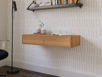 A minimalist room showcases a handmade aesthetic with an American Cherry wall-mounted shelf displaying a potted plant and books, enhanced by the wall’s subtle linear patterns. On the left, you can partially see a black floor lamp next to the Krovel Floating Desk in White Oak and a floating desk chair.