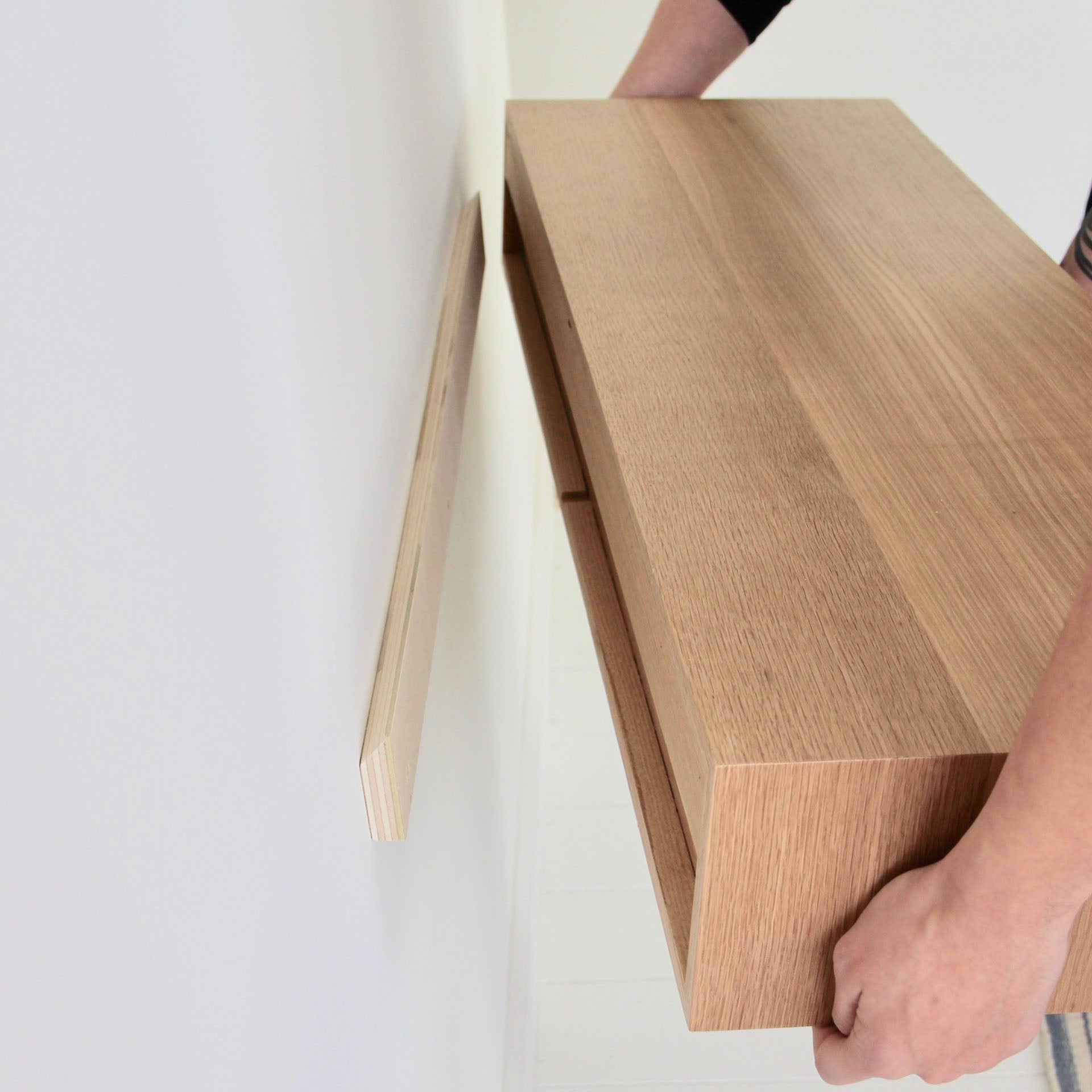 A person is mounting a Krovel Floating Desk in White Oak onto a wall. The hands carefully align the desk with a bracket on the plain white wall, showcasing its natural wood finish.