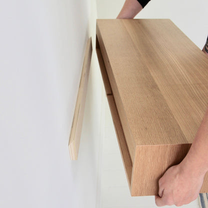 A person is mounting a Krovel Floating Desk in White Oak onto a wall. The hands carefully align the desk with a bracket on the plain white wall, showcasing its natural wood finish.