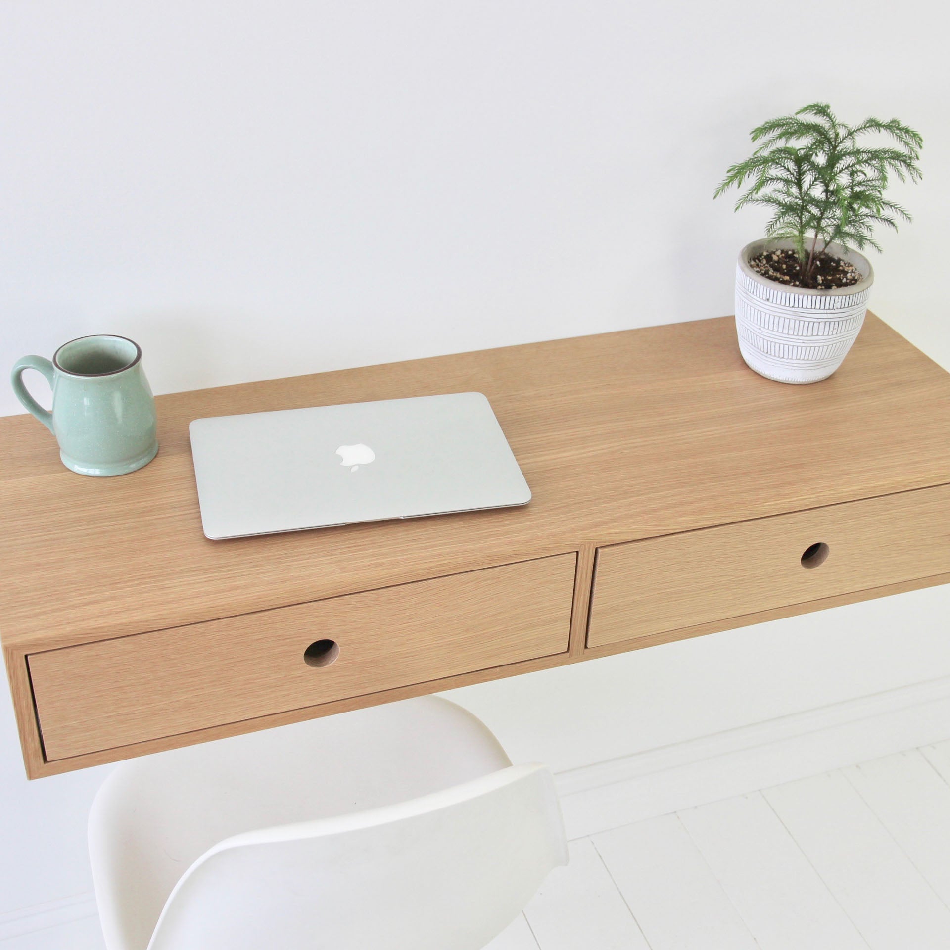 A minimalist handmade Floating Desk in White Oak by Krovel, featuring two drawers, supports a closed silver laptop and a green mug. A small potted plant sits on the corner, while a white chair is nearby. The background is a serene white wall and floor.