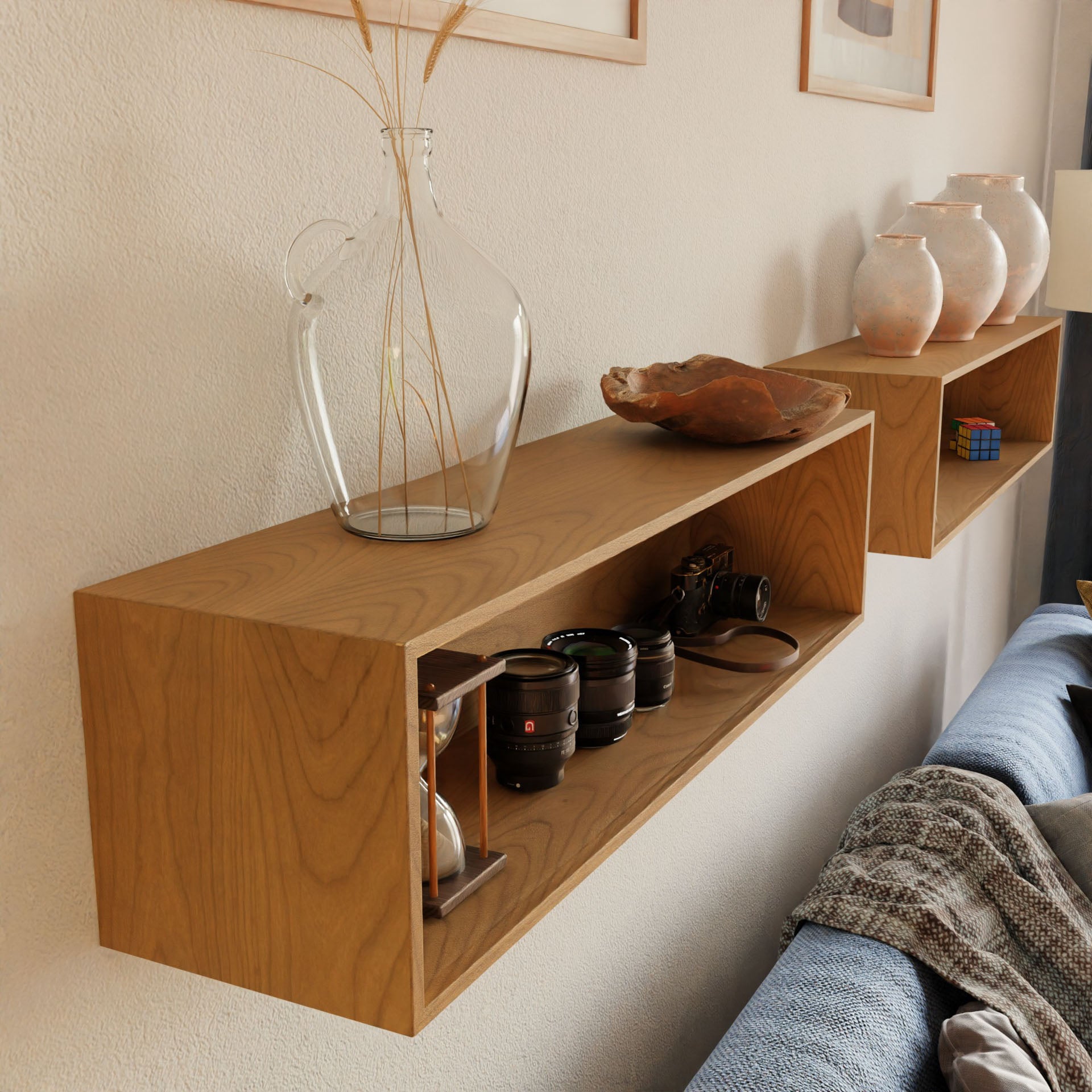 A Krovel Floating Display Case in Cherry showcases glass jugs, decorative pottery, and several camera lenses alongside a camera. Below the case is a partially visible blue couch with a patterned blanket, highlighting the sleek elegance of its French cleat design.