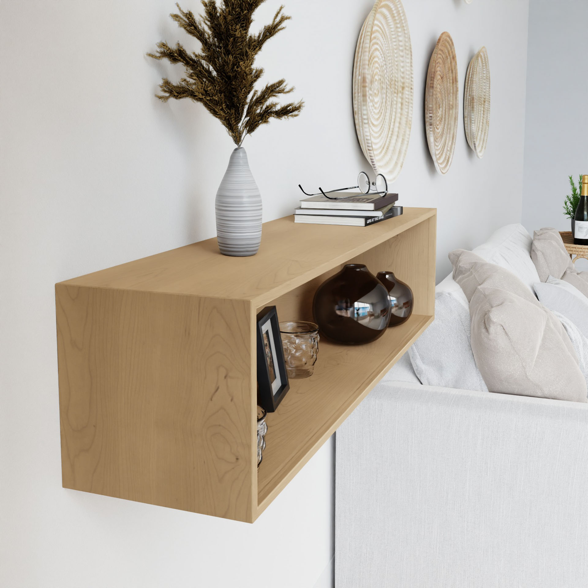 A Krovel Floating Display Case in Maple, securely mounted on a white wall with a French cleat, elegantly showcases a white vase filled with dried plants, alongside books, glasses, a framed photo, and two amber glass vases. Above the light gray sofa hang textured circular art pieces.